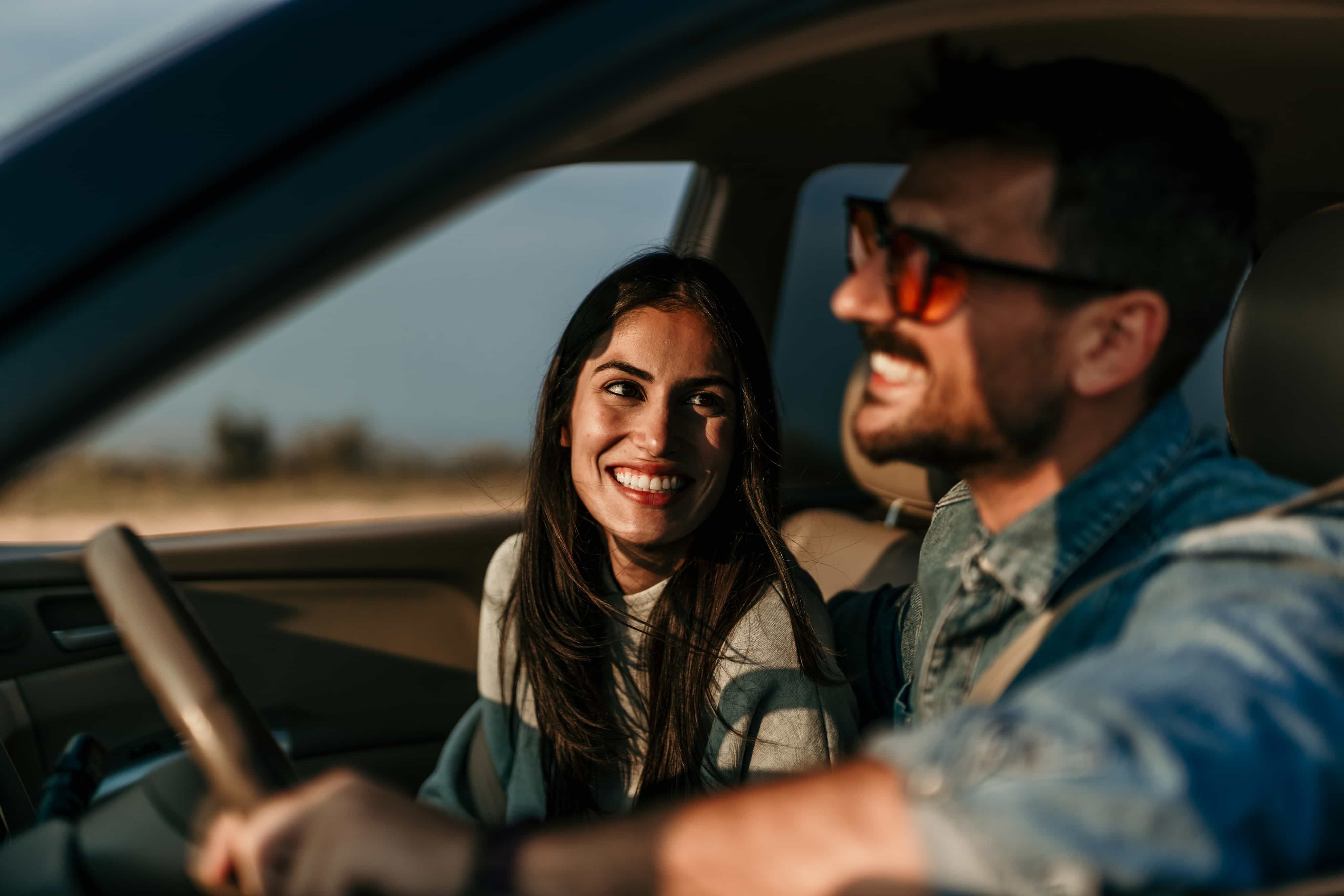 A woman and man smiling in a car, enjoying a sunny drive. The woman looks at him while he focuses on the road, wearing sunglasses.