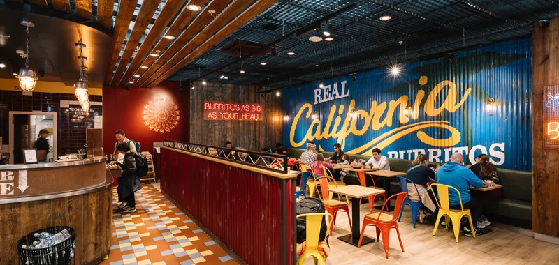 Colourful restaurant with wooden décor, neon signs, and diners seated at tables. Large "Real California Burritos" mural on the wall.