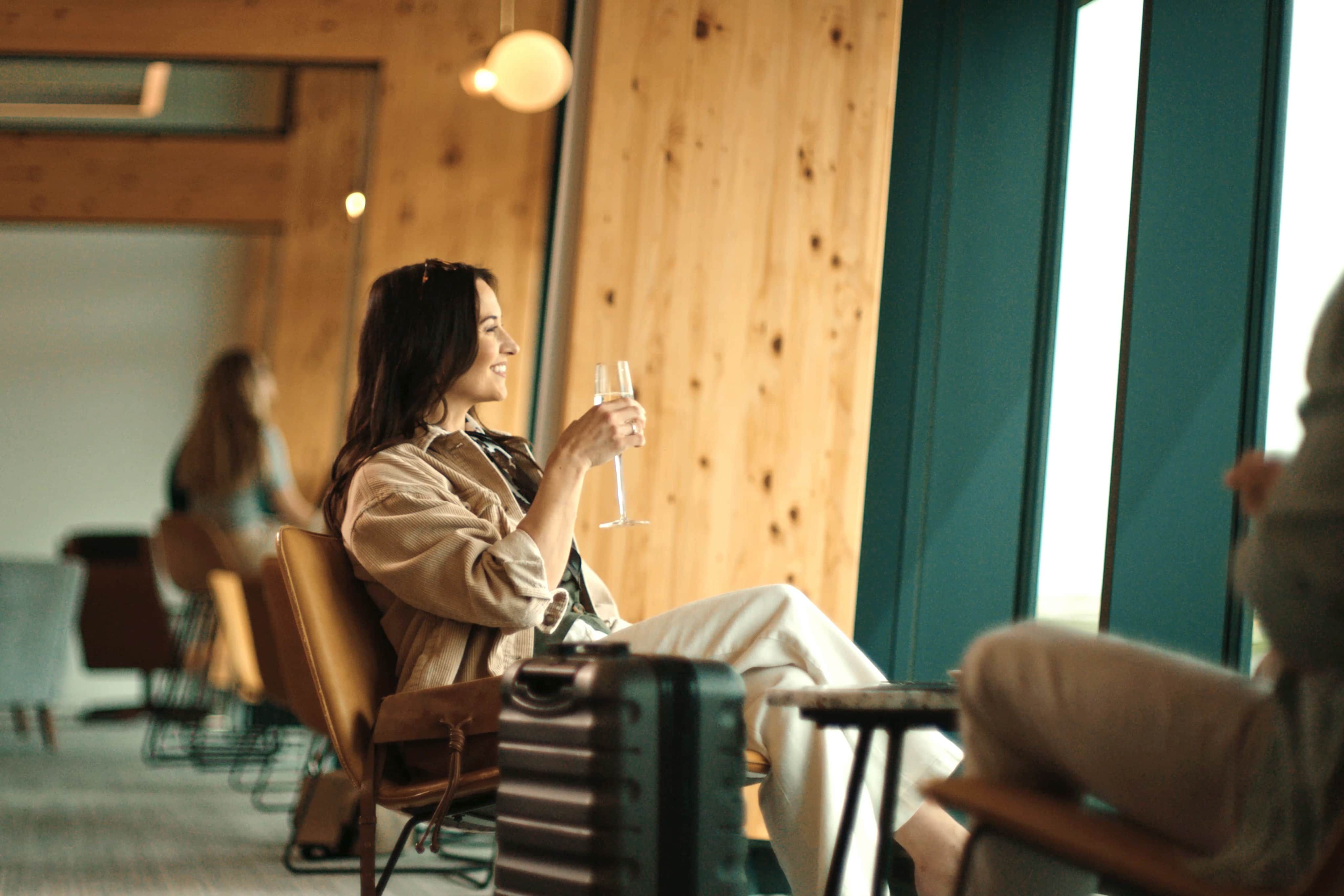 Woman relaxing with a drink by large windows in a modern, wooden interior, with luggage nearby.