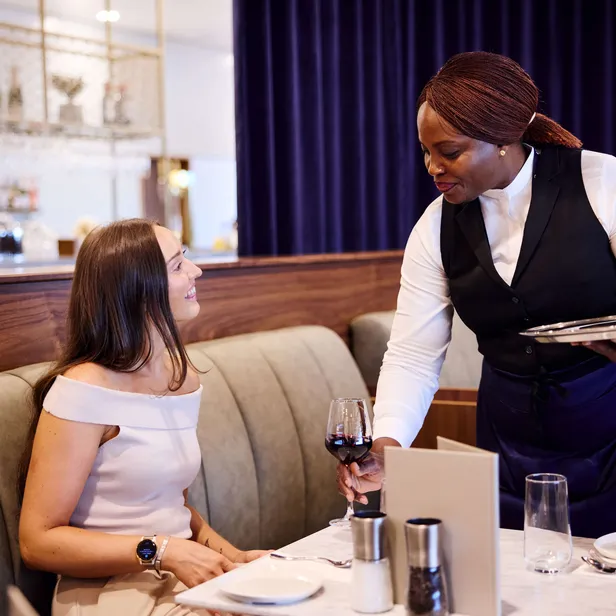 A smiling woman sits at a restaurant table while a server hands her a glass of red wine.