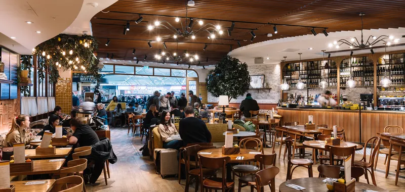 Cosy restaurant interior with warm lighting, wooden tables, passengers dining, and a stocked bar against a backdrop of decorative plants and ambient décor.