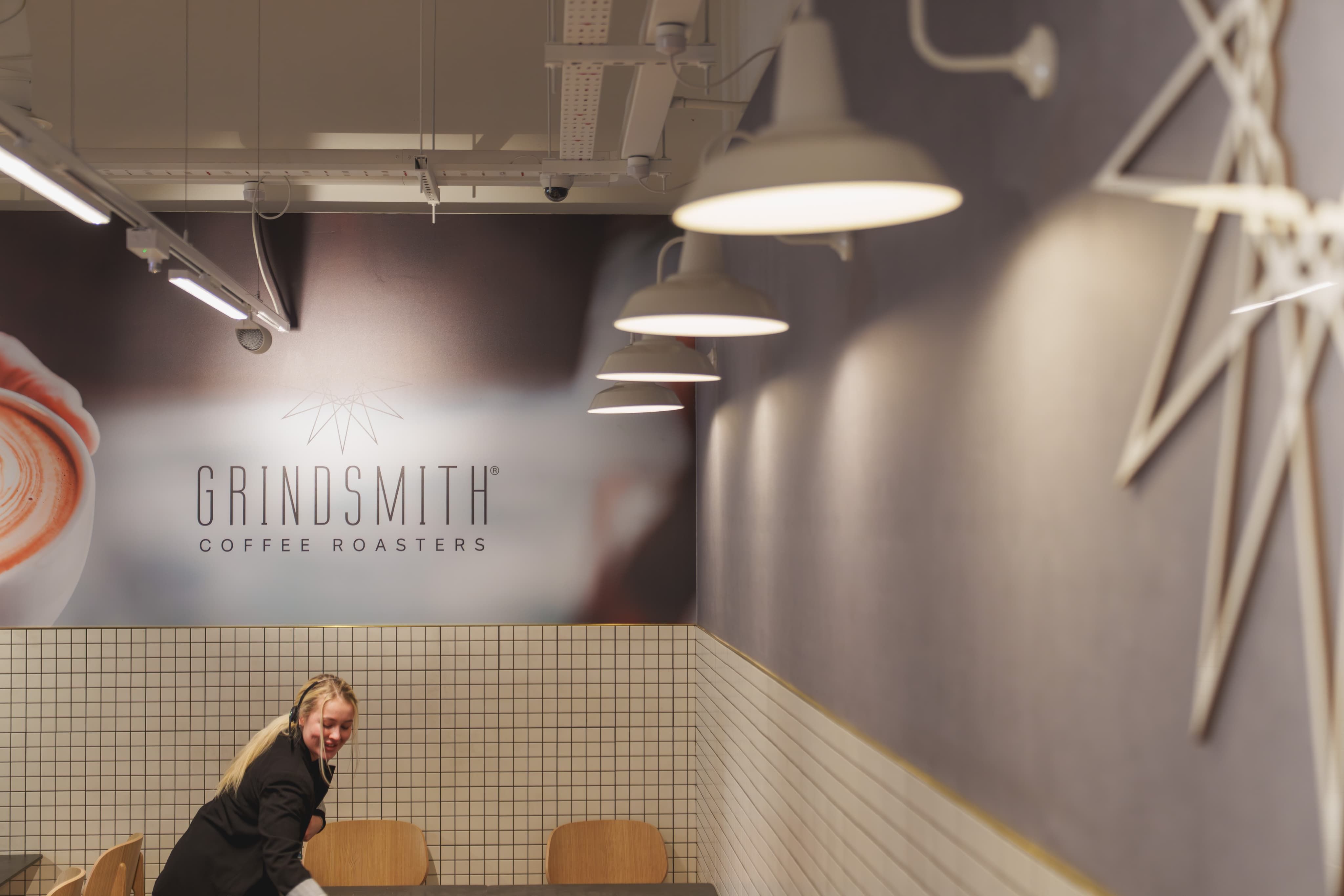 A person arranging a table in a modern coffee shop with a "Grindsmith Coffee Roasters" sign on the wall and white hanging lights.