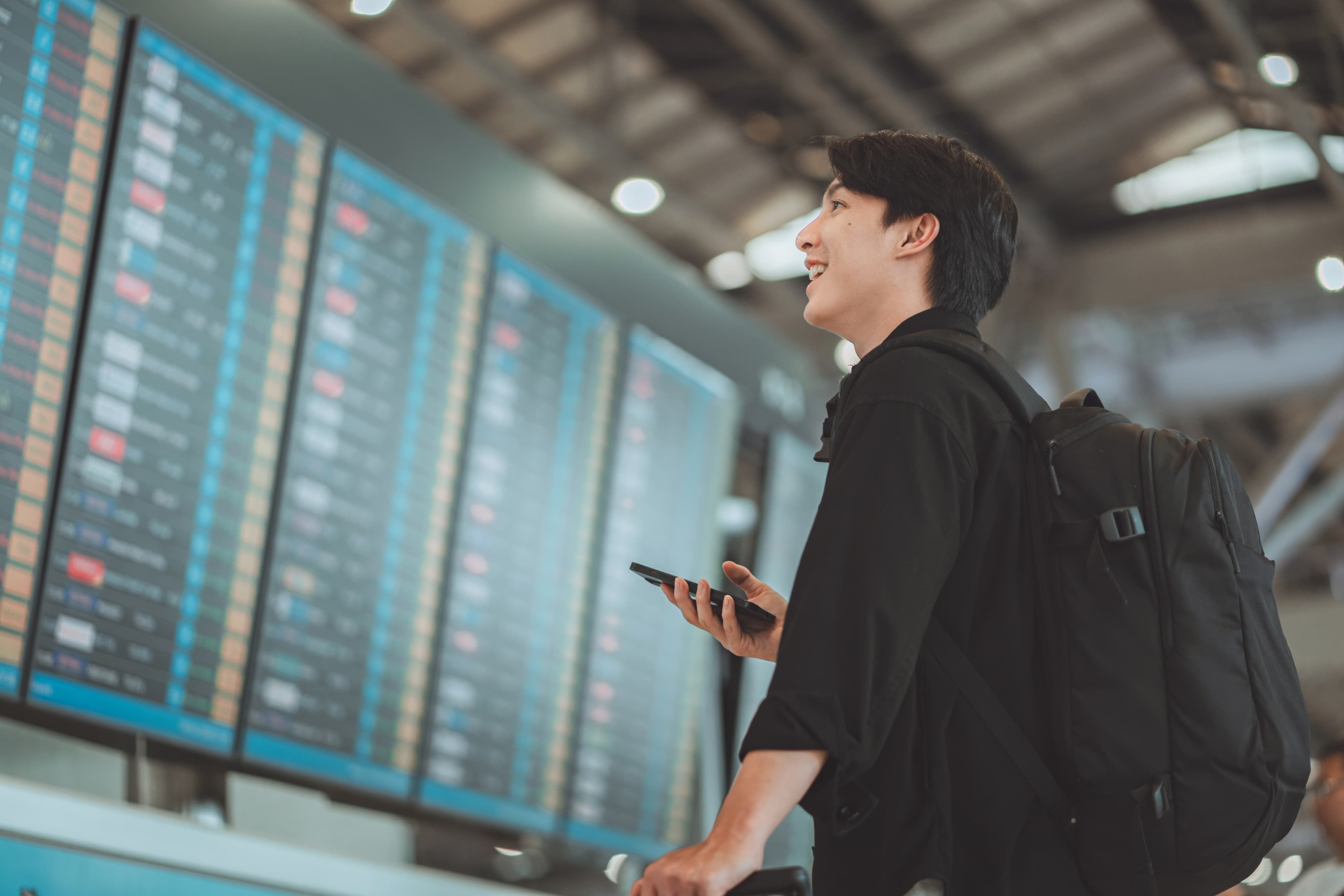 A person with a backpack holds a smartphone and looks up at a large airport departures board displaying various flight schedules.