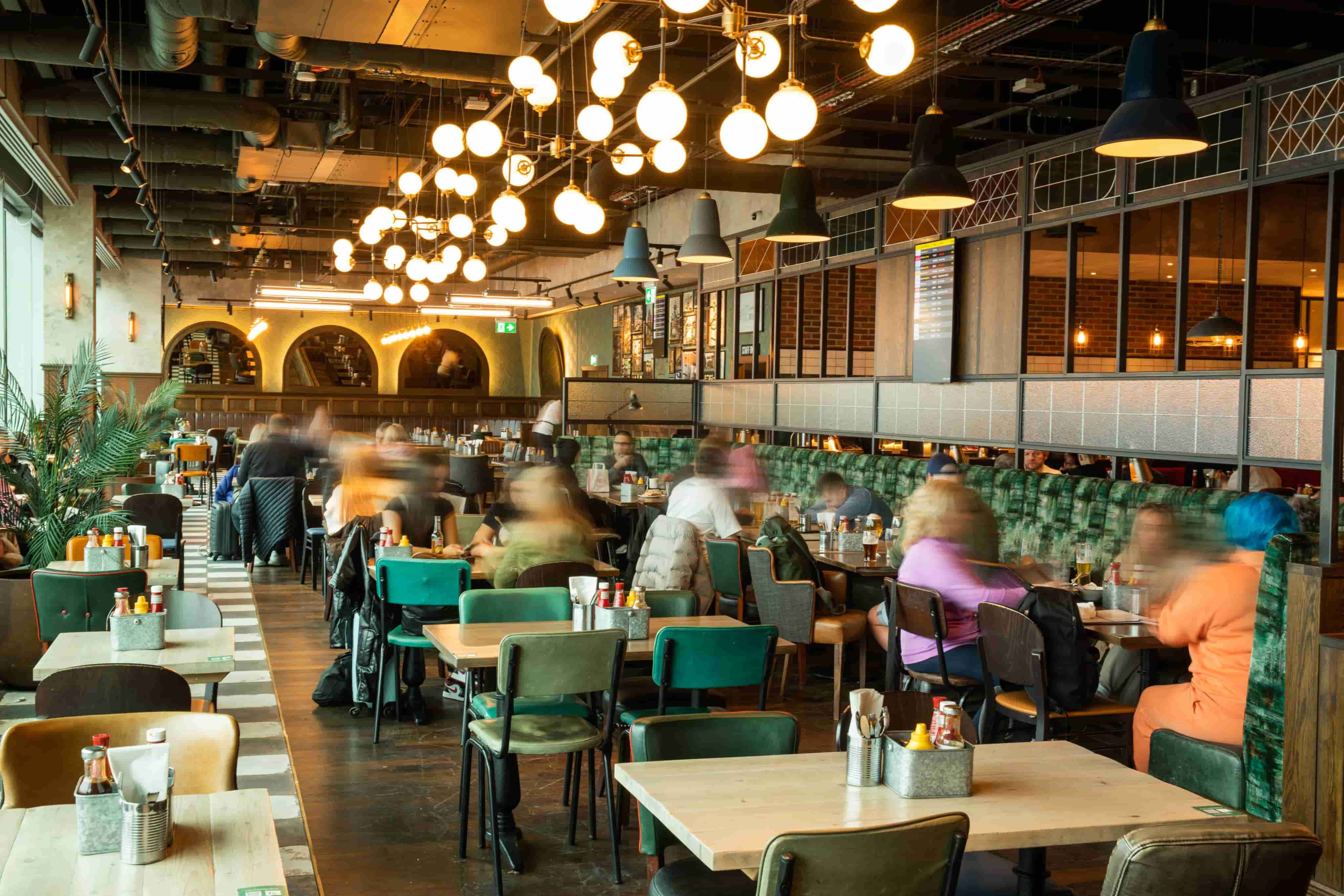 A bustling restaurant with modern decor, featuring wooden tables, green chairs, and warm lighting. Diners create a lively atmosphere.