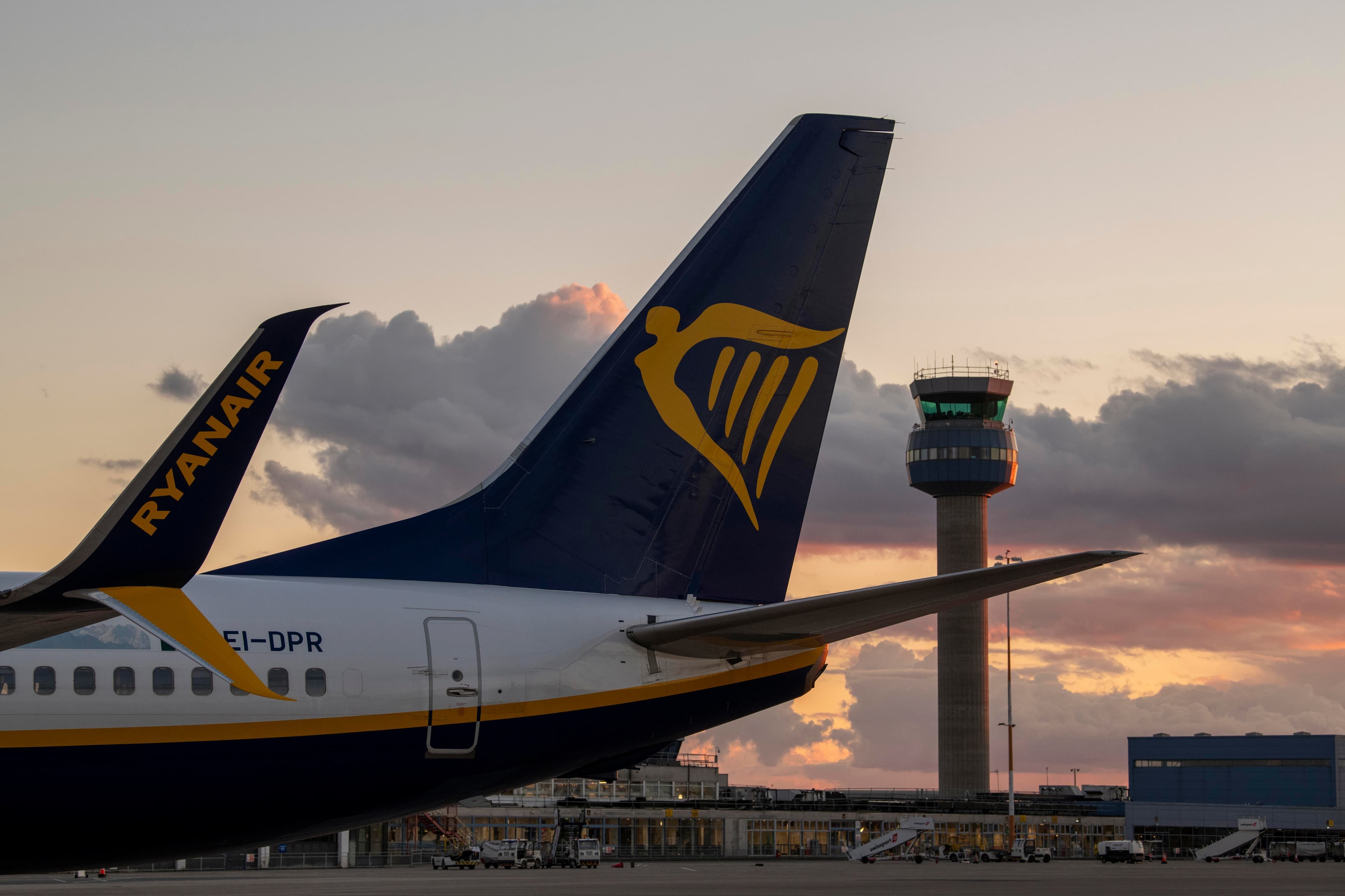 Ryanair plane tail with logo at sunset, airport control tower in background, cloudy sky.