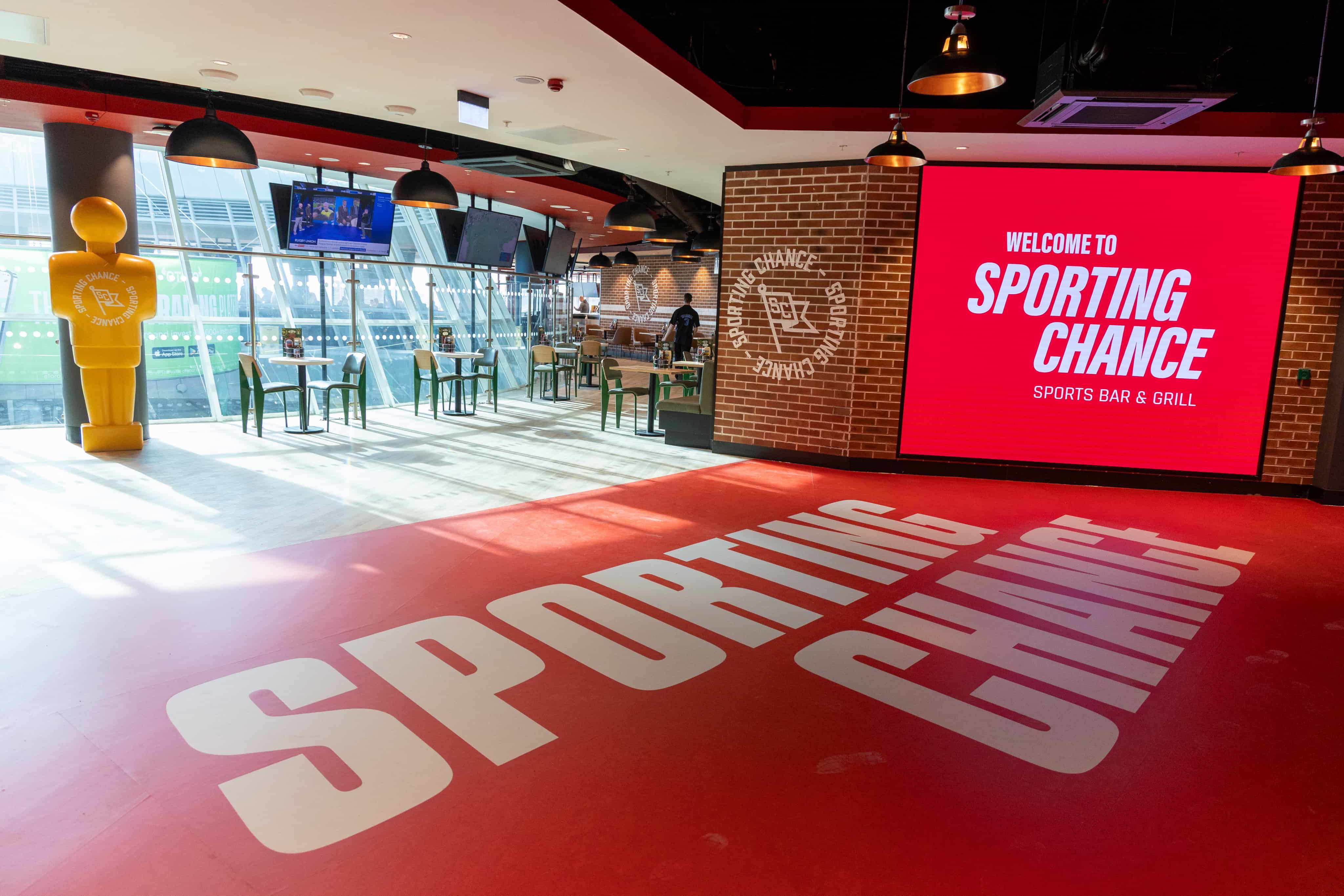 Spacious sports bar with large red flooring, tables, a brick wall, and a big screen displaying "Welcome to Sporting Chance Sports Bar & Grill."