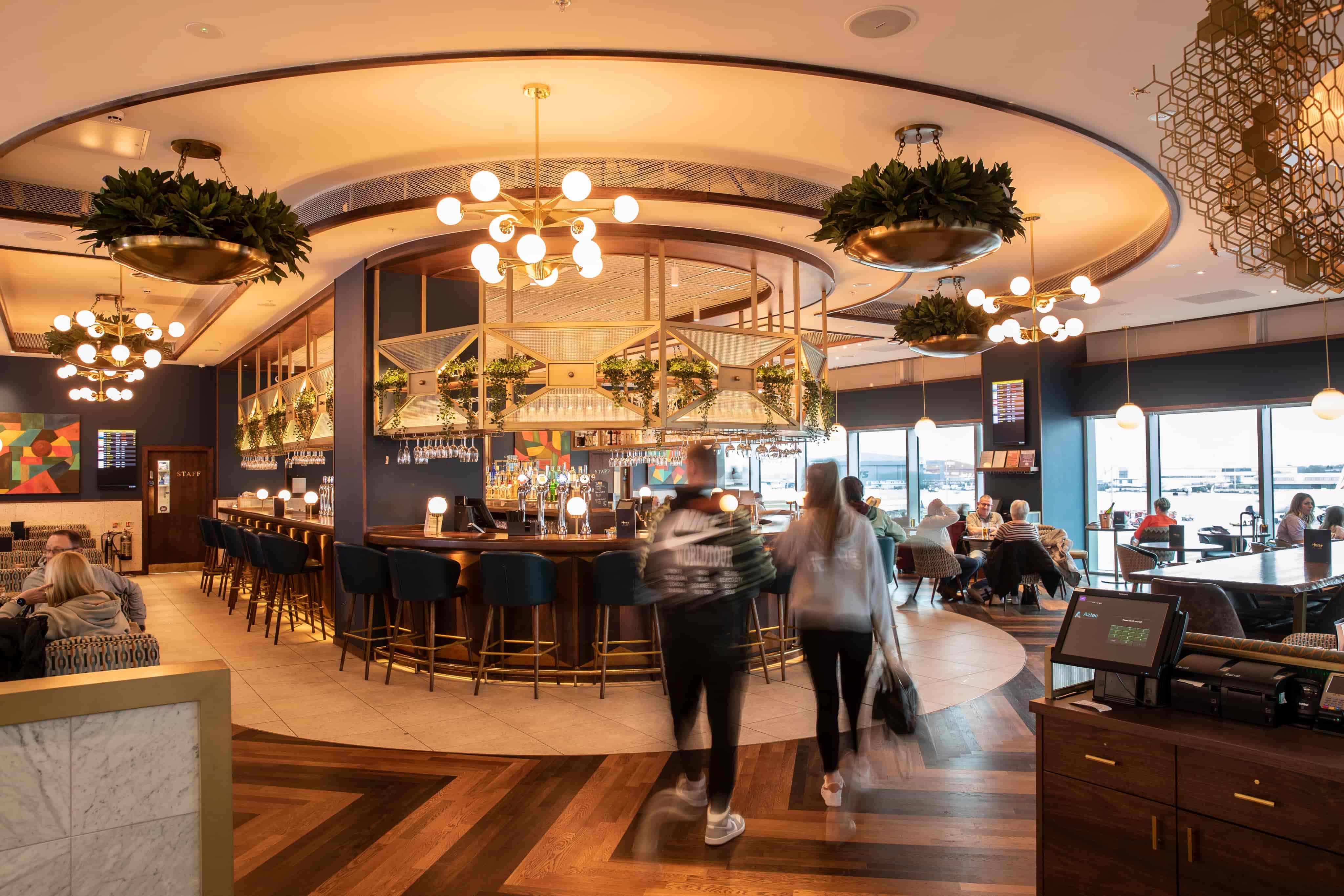 Modern airport lounge with bar seating, suspended greenery, and customers. Floor-to-ceiling windows provide views of the runway.