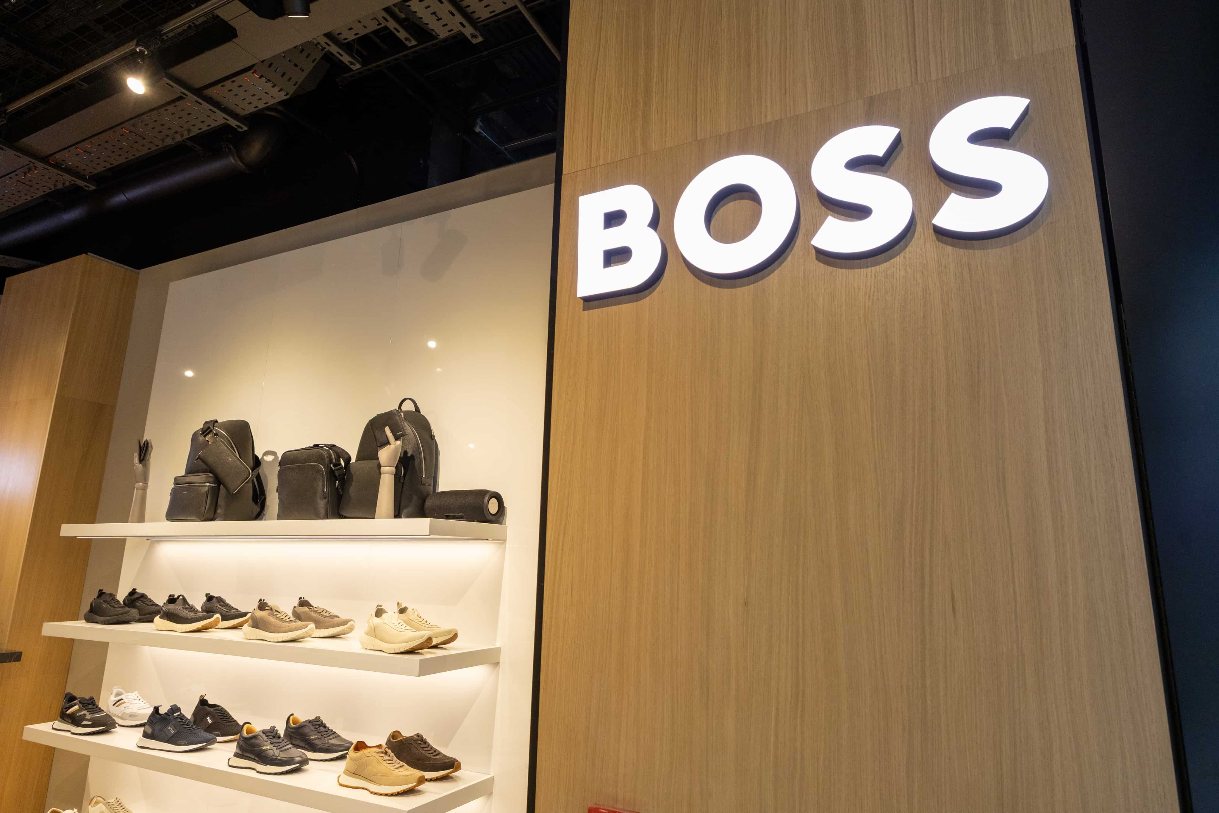 Store display featuring shelves with various sneakers and bags. The word "BOSS" is illuminated on the wooden wall above.