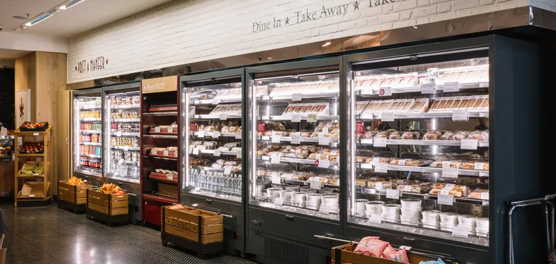 A well-lit refrigerated display in a store with sandwiches, drinks, and packaged foods. Wooden crates hold fresh fruit and snacks.