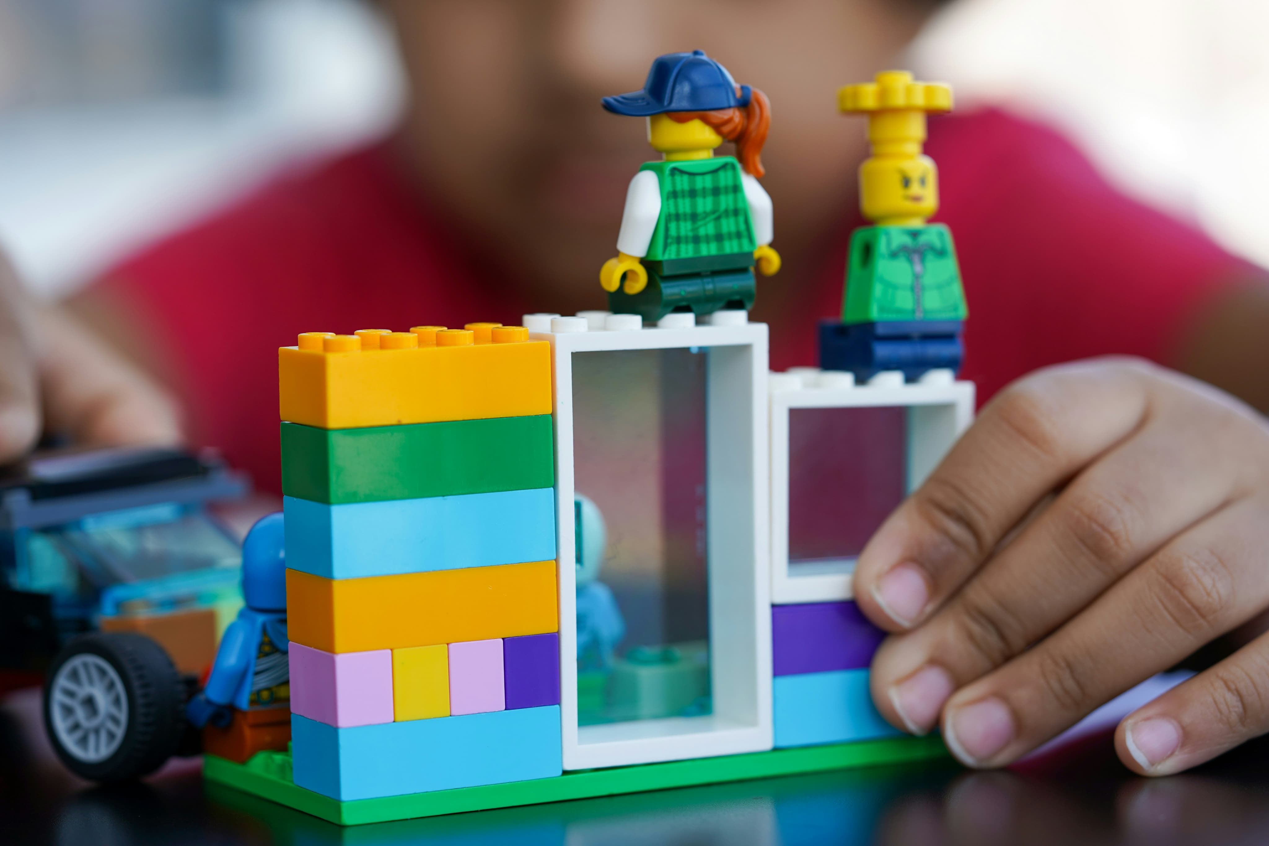 Child's hands assembling a colorful LEGO building with stacked bricks and two minifigures on top.