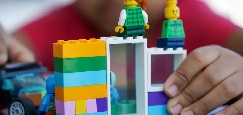 Child's hands assembling a colorful LEGO building with stacked bricks and two minifigures on top.