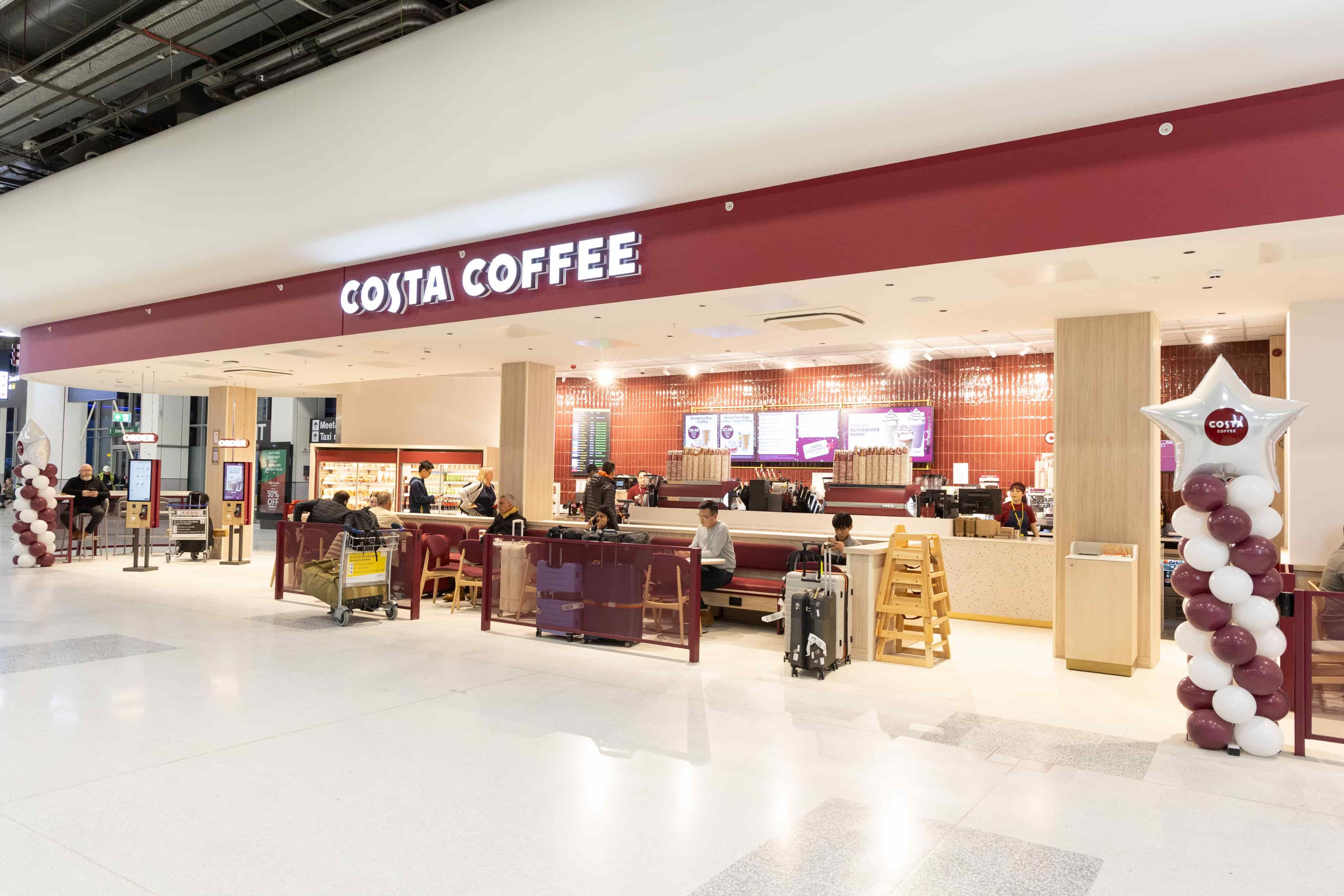 Wide view of a Costa Coffee shop inside an airport, with people seated and ordering. Decor includes red accents and balloon arrangements.