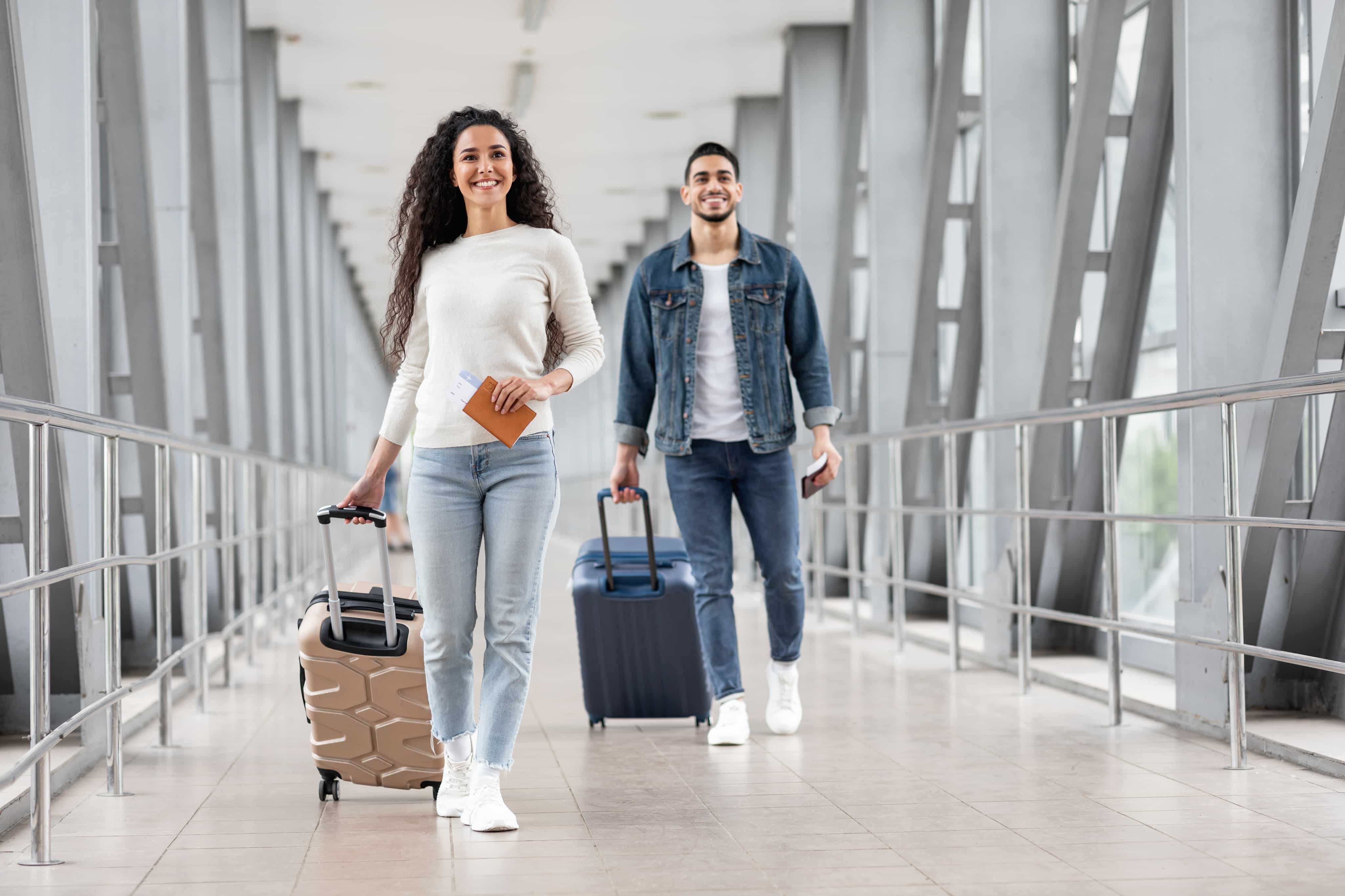 Two people walking through an airport corridor with luggage and passports, smiling and dressed casually in jeans and light-colored tops.