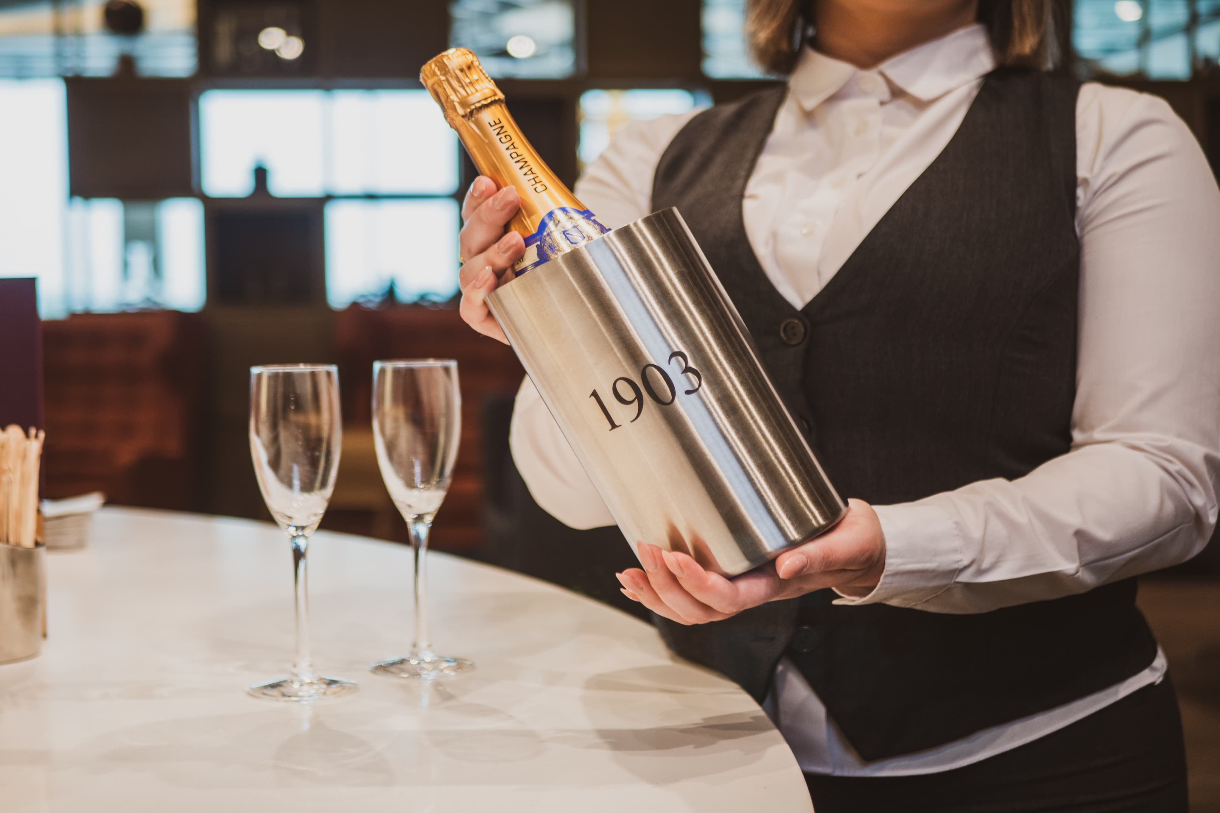 Server presenting a bottle of champagne in a bucket labelled "1903," with two empty champagne flutes on a nearby table.