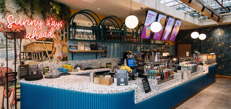 A modern café with a colourful counter and neon sign reading "Sunny days ahead." Hanging lights, menu screens, and tropical-themed decor are visible.