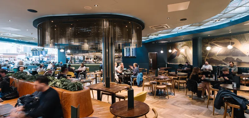 Spacious café with modern décor, wooden tables, and customers dining and socialising. Wall art depicts birds.