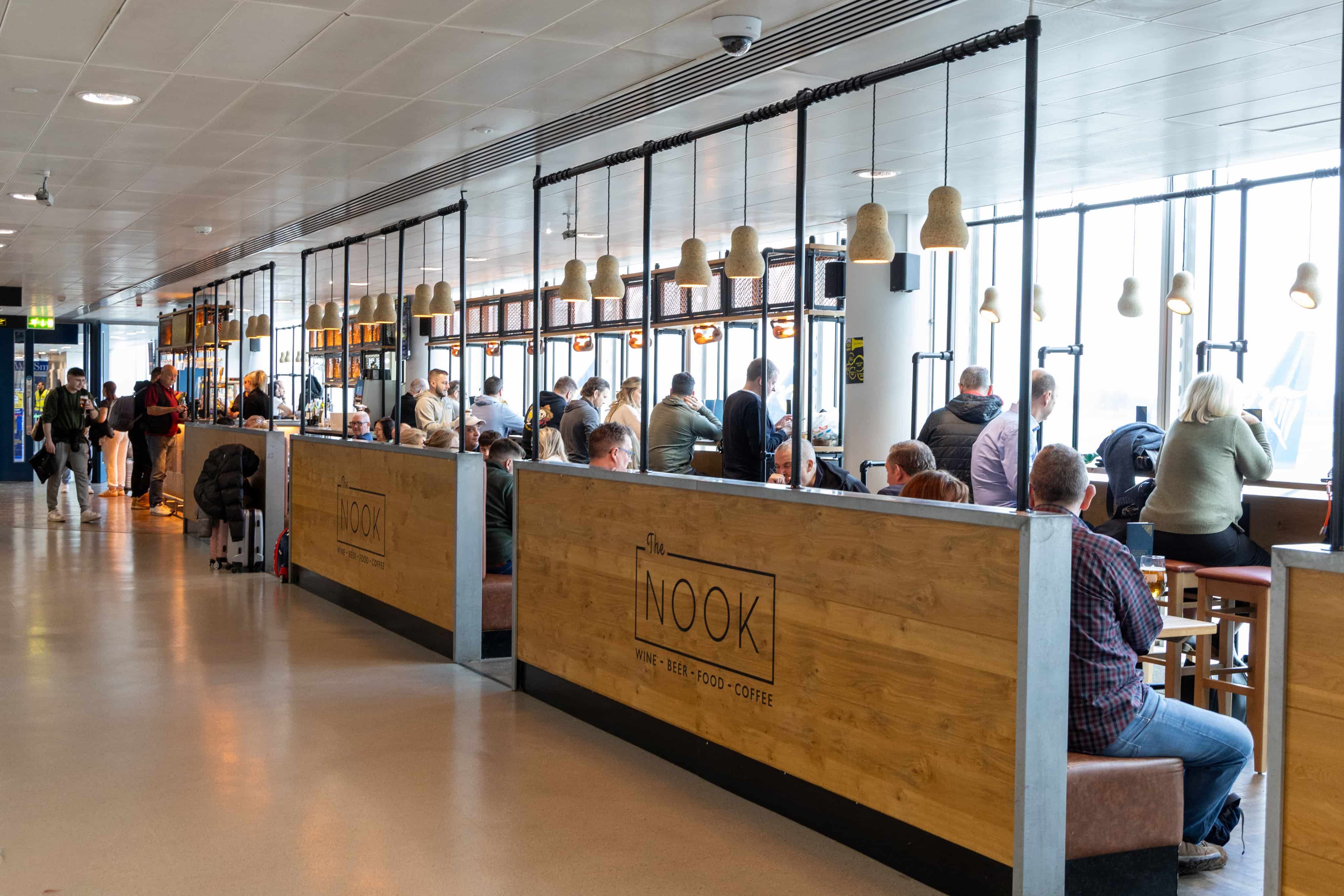People seated at wooden tables inside an airport cafe, with overhead lamps and large windows in the background.