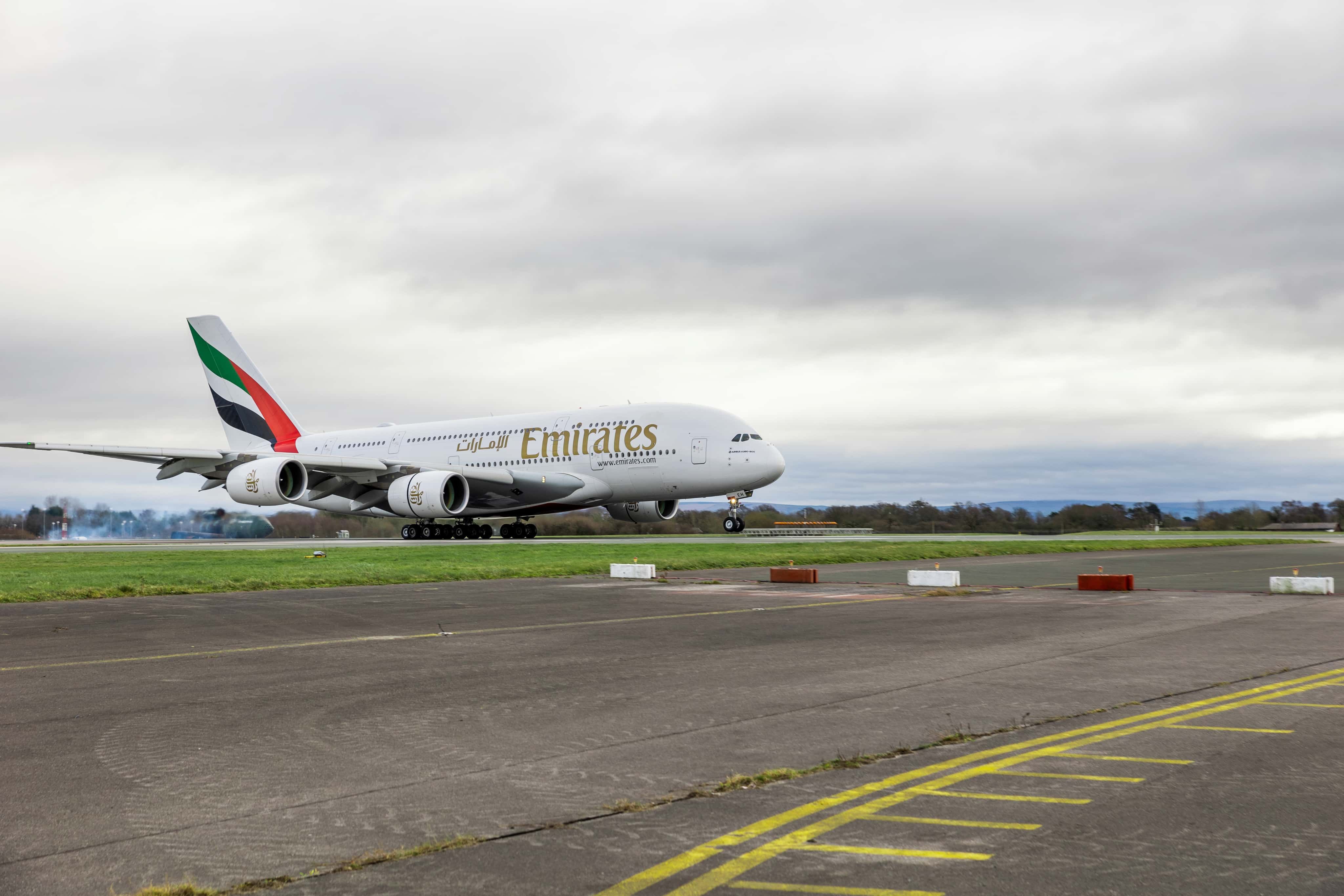 An Emirates Airbus A380 lands on a runway surrounded by grass, under a cloudy sky.