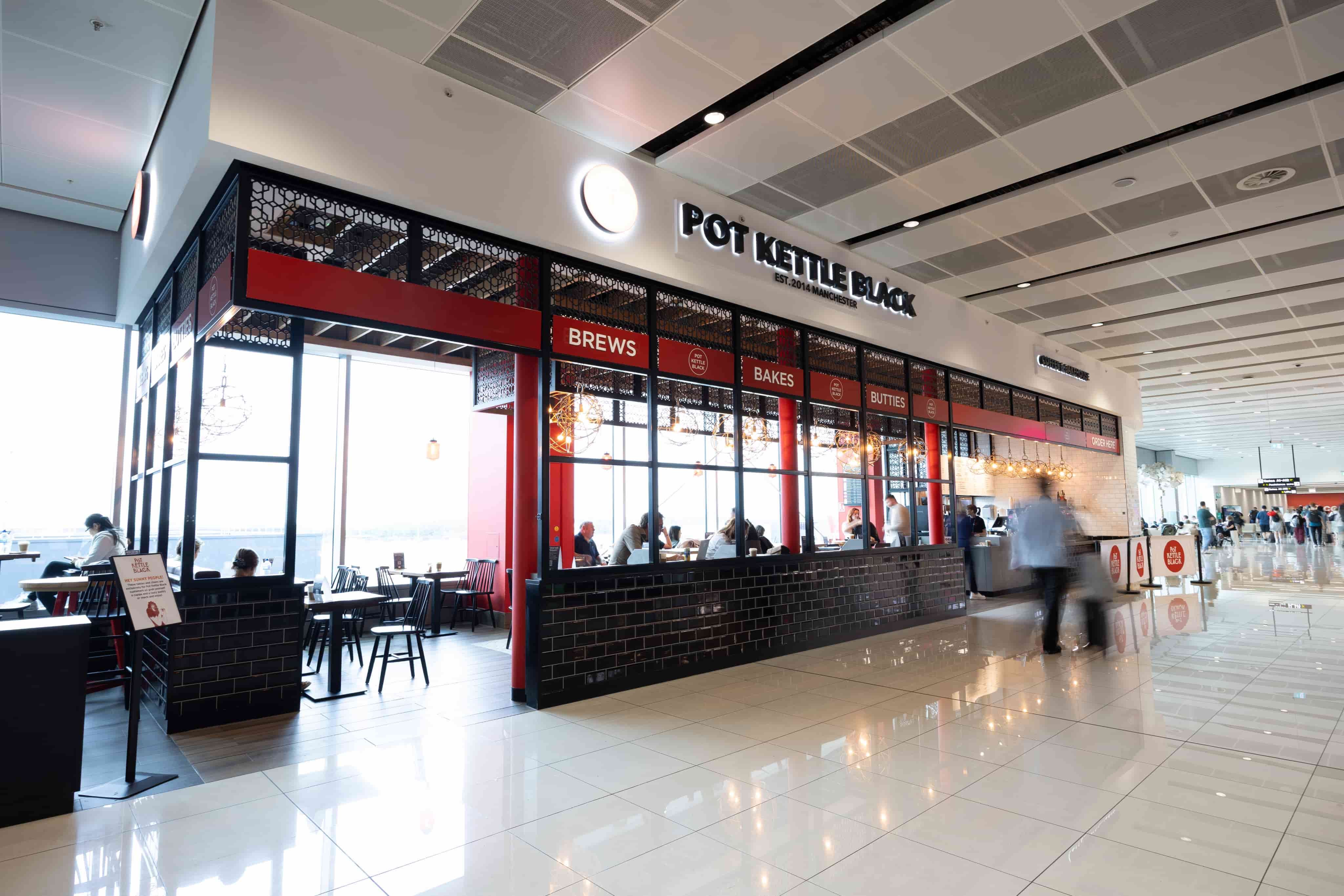Modern airport café with large glass windows, black and red accents, and a sign reading "Pot Kettle Black." People are walking nearby.