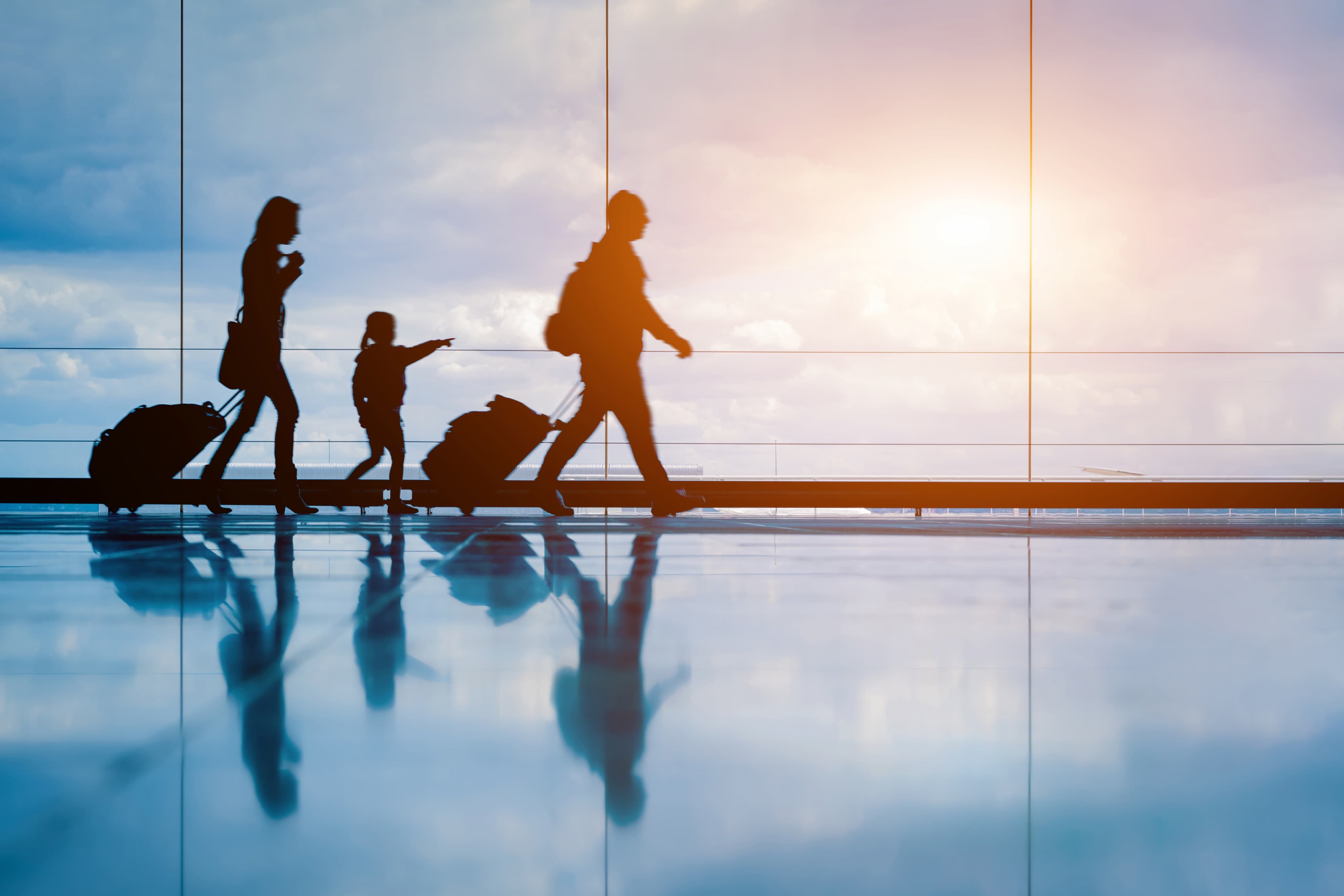 Silhouettes of a family with luggage walking through an airport terminal, with a bright sunset visible through large windows.