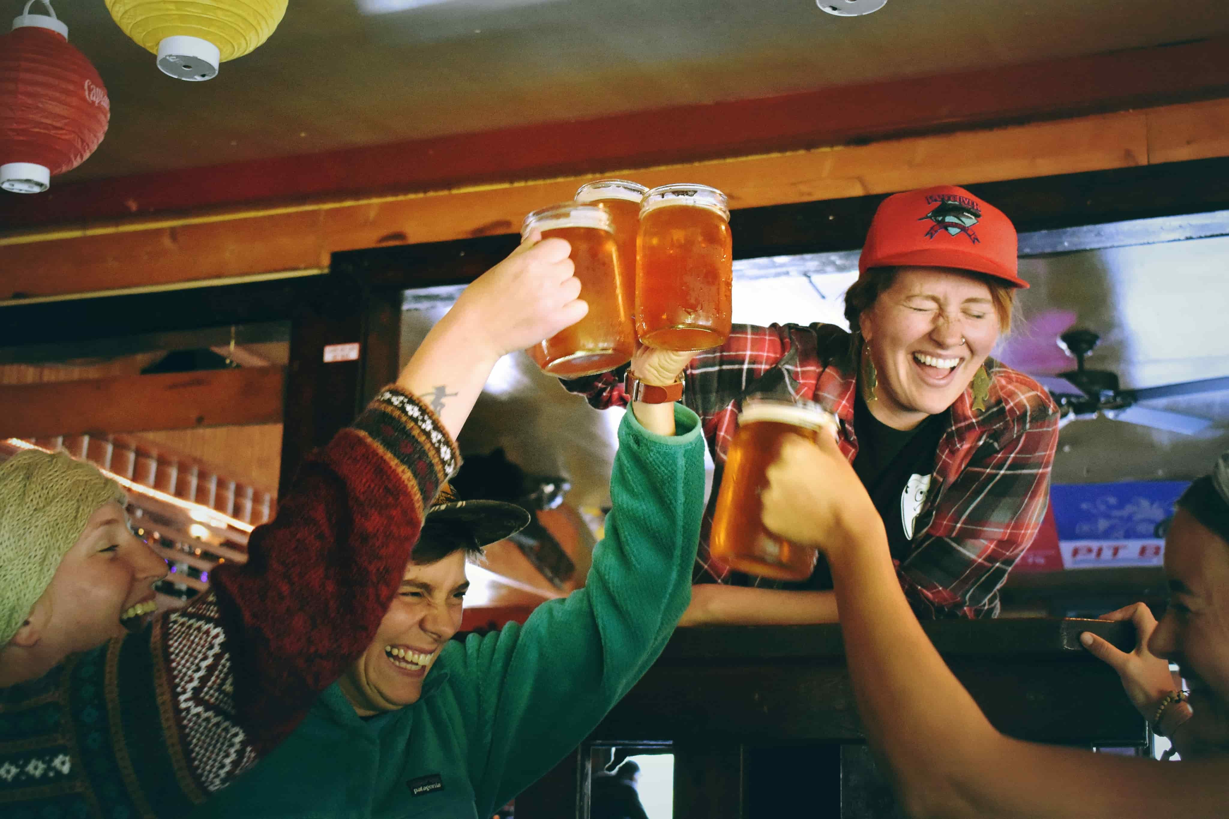 Four people cheerfully clinking beer glasses in a cozy pub setting, with warm lighting and colorful decor.