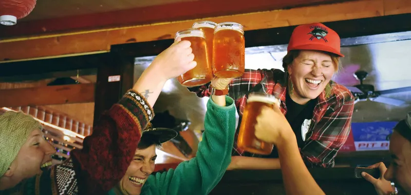 Four people cheerfully clinking beer glasses in a cozy pub setting, with warm lighting and colorful decor.