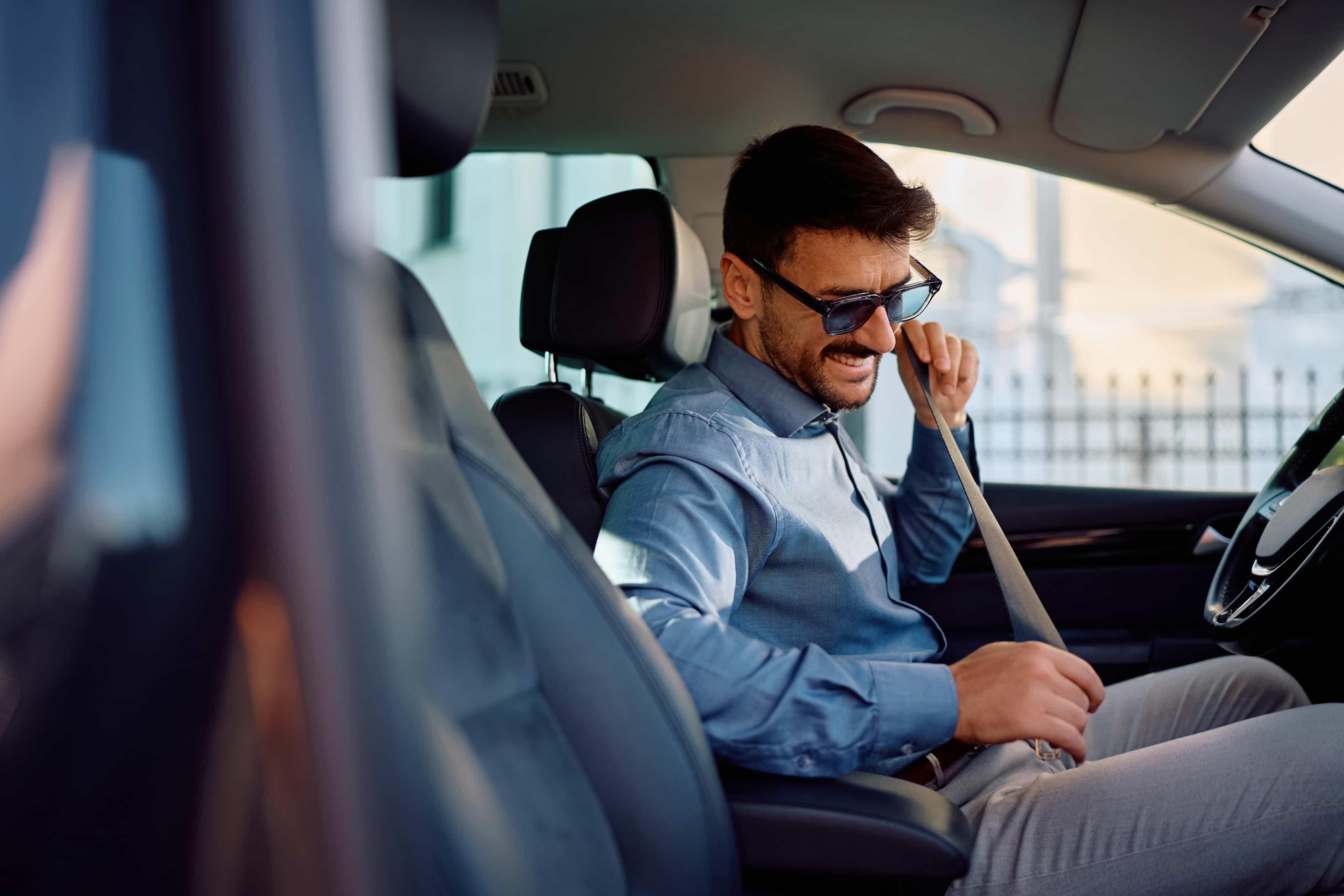 Man in sunglasses fastens seatbelt while sitting inside a car, wearing a blue shirt and smiling.