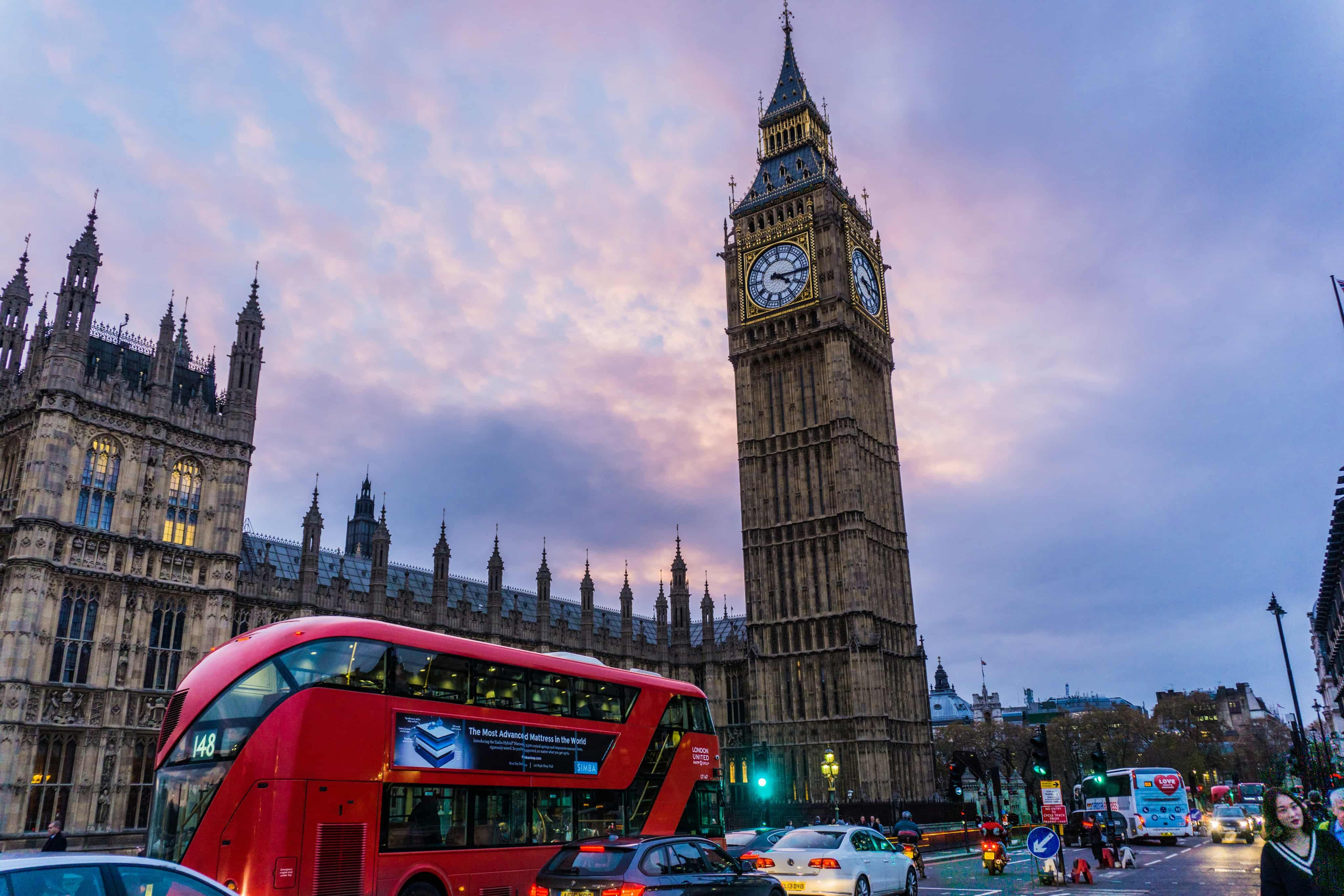 Big Ben at dusk with a red double-decker bus and cars in the foreground, under a colorful sky.