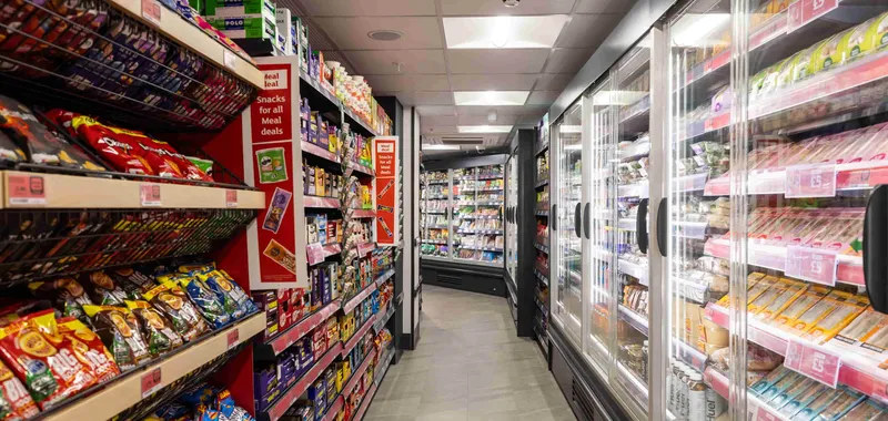 Grocery store aisle with shelves of snacks on the left and refrigerated food items on the right, under bright overhead lights.