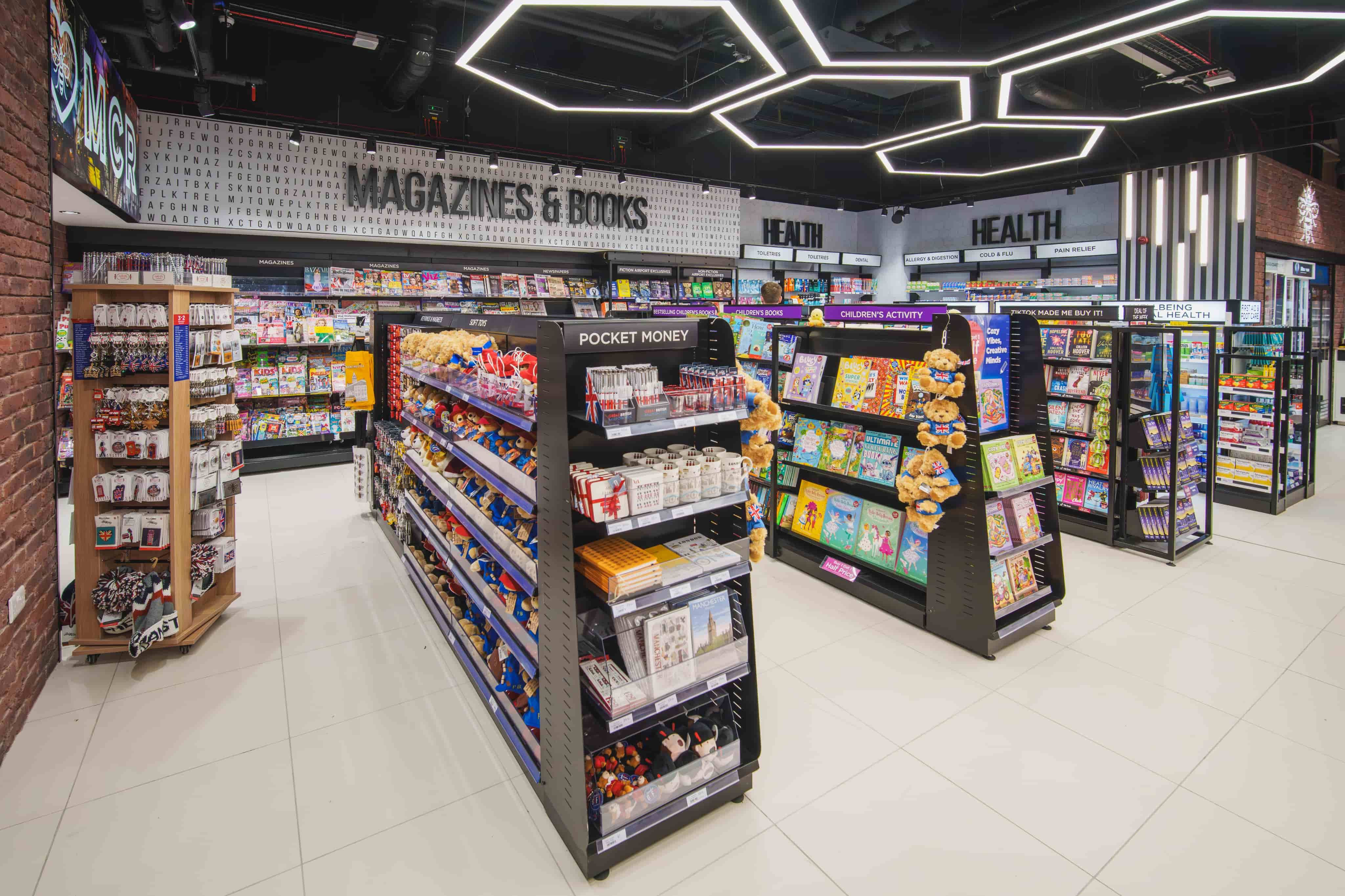 A well-lit bookstore with colorful shelves displaying magazines, books, and health products. Modern ceiling lights illuminate the space.