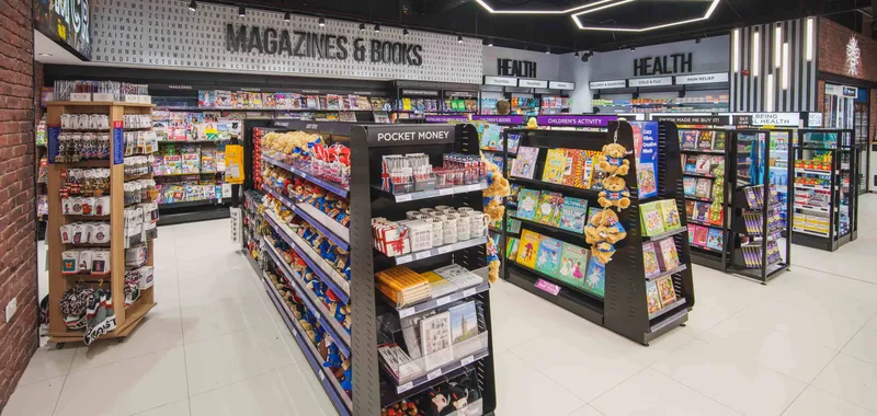 A well-lit bookstore with colorful shelves displaying magazines, books, and health products. Modern ceiling lights illuminate the space.
