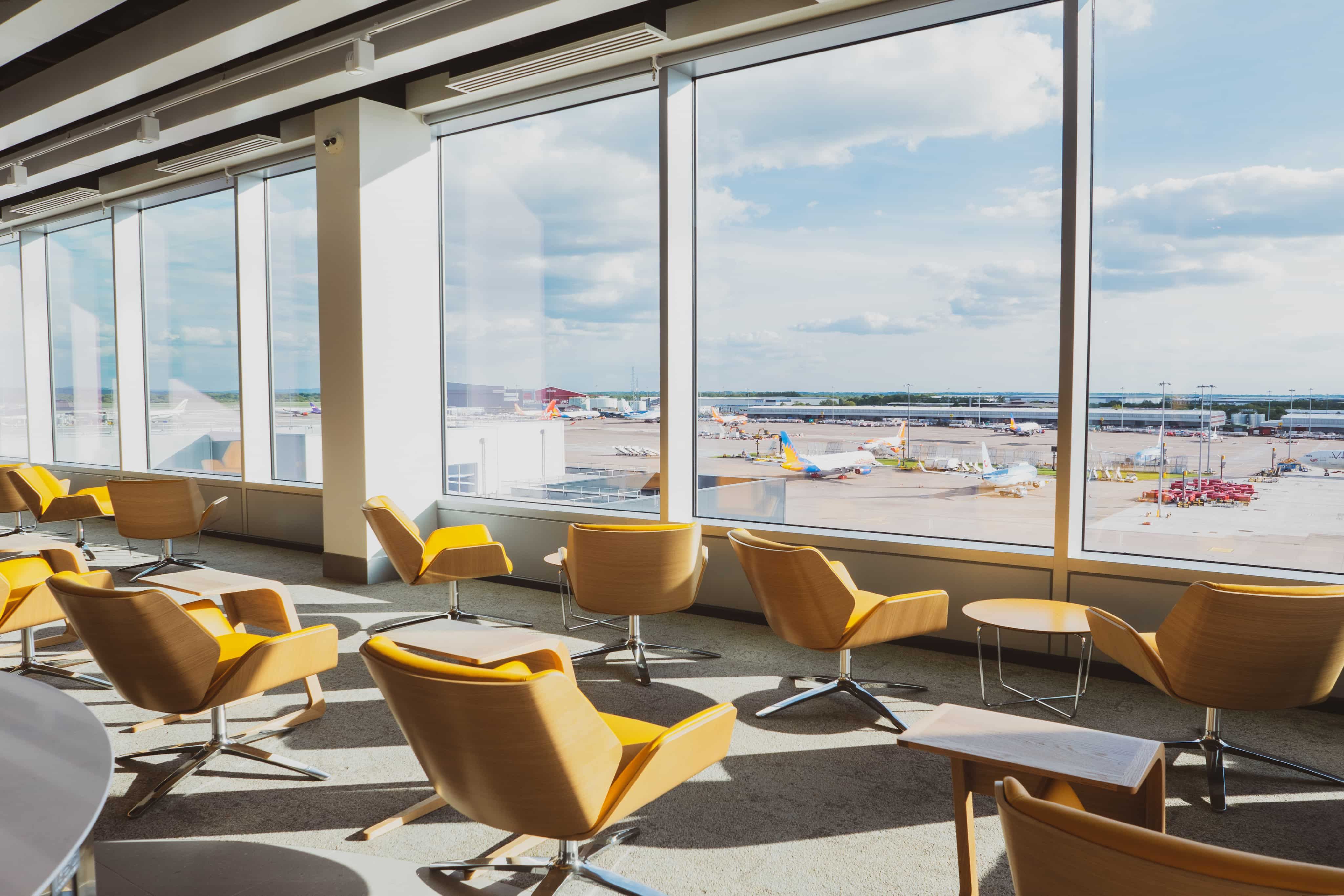 Sunlit airport lounge with yellow chairs facing large windows, overlooking a busy airport runway with several airplanes under a clear blue sky.