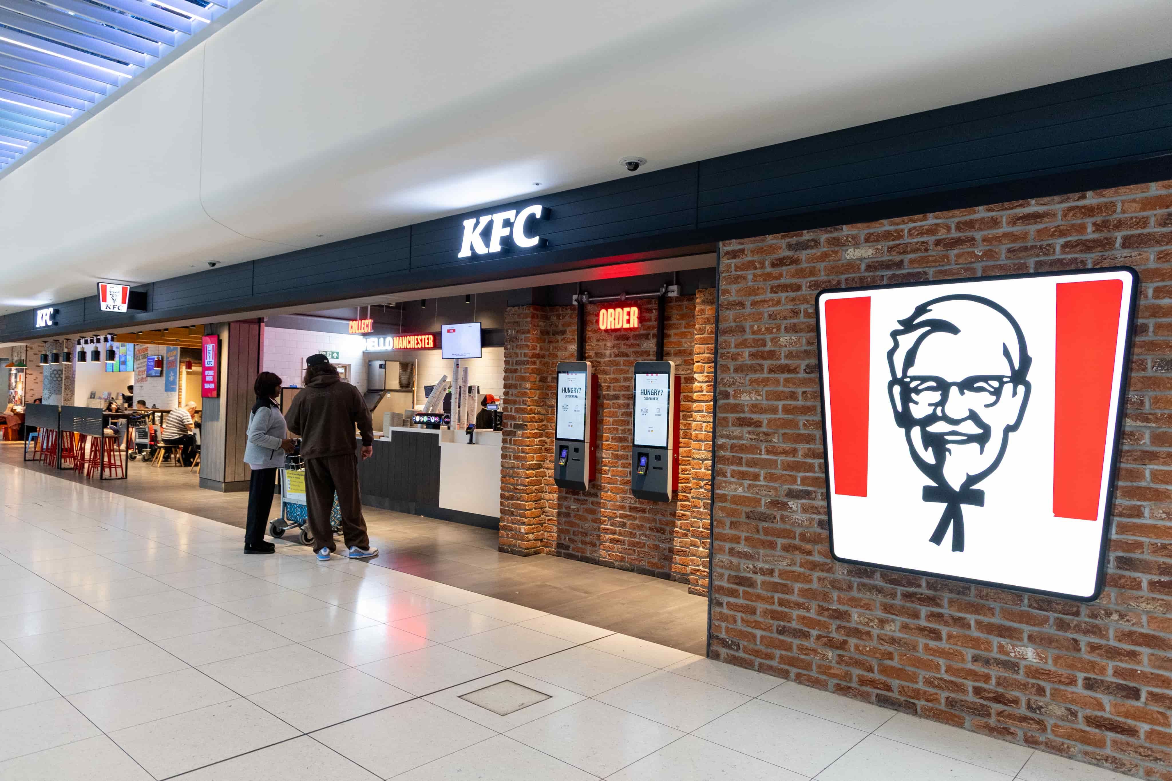 KFC restaurant interior with digital ordering kiosks, people standing in line, and a prominent logo on a brick wall.
