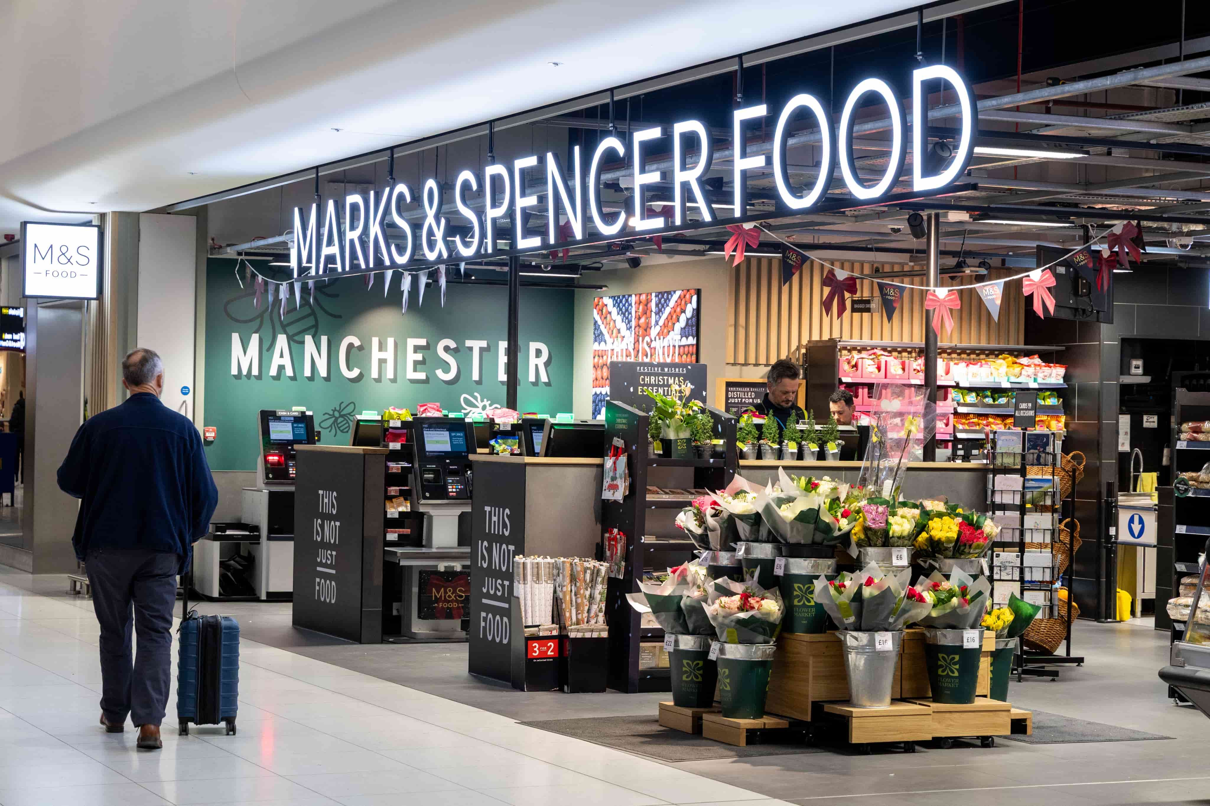 A traveler with a suitcase approaches a brightly lit Marks & Spencer Food store in Manchester, featuring a flower display and self-checkouts.