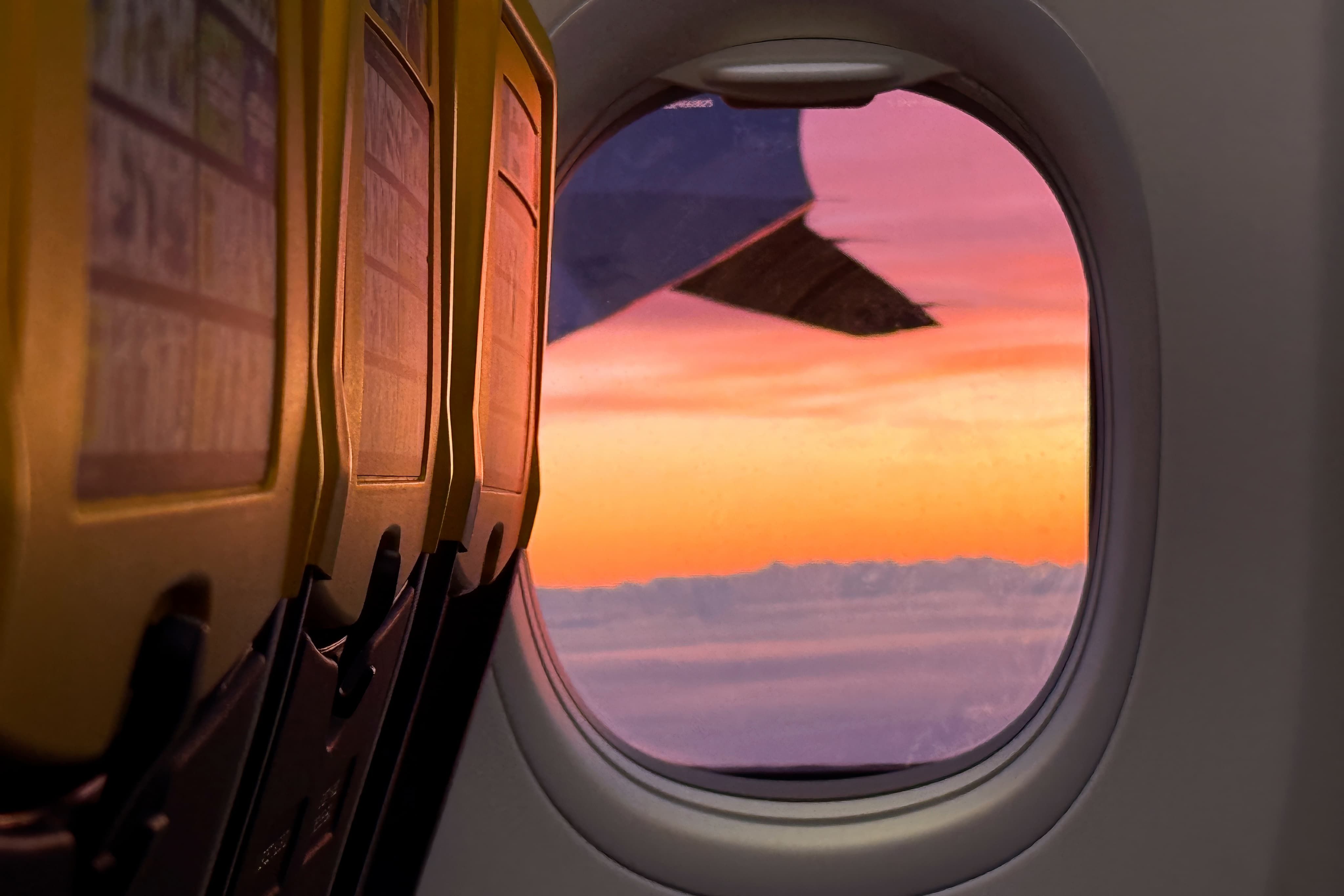 Airplane window view with a vibrant pink and orange sunset over the clouds, seats with yellow backs visible on the left.