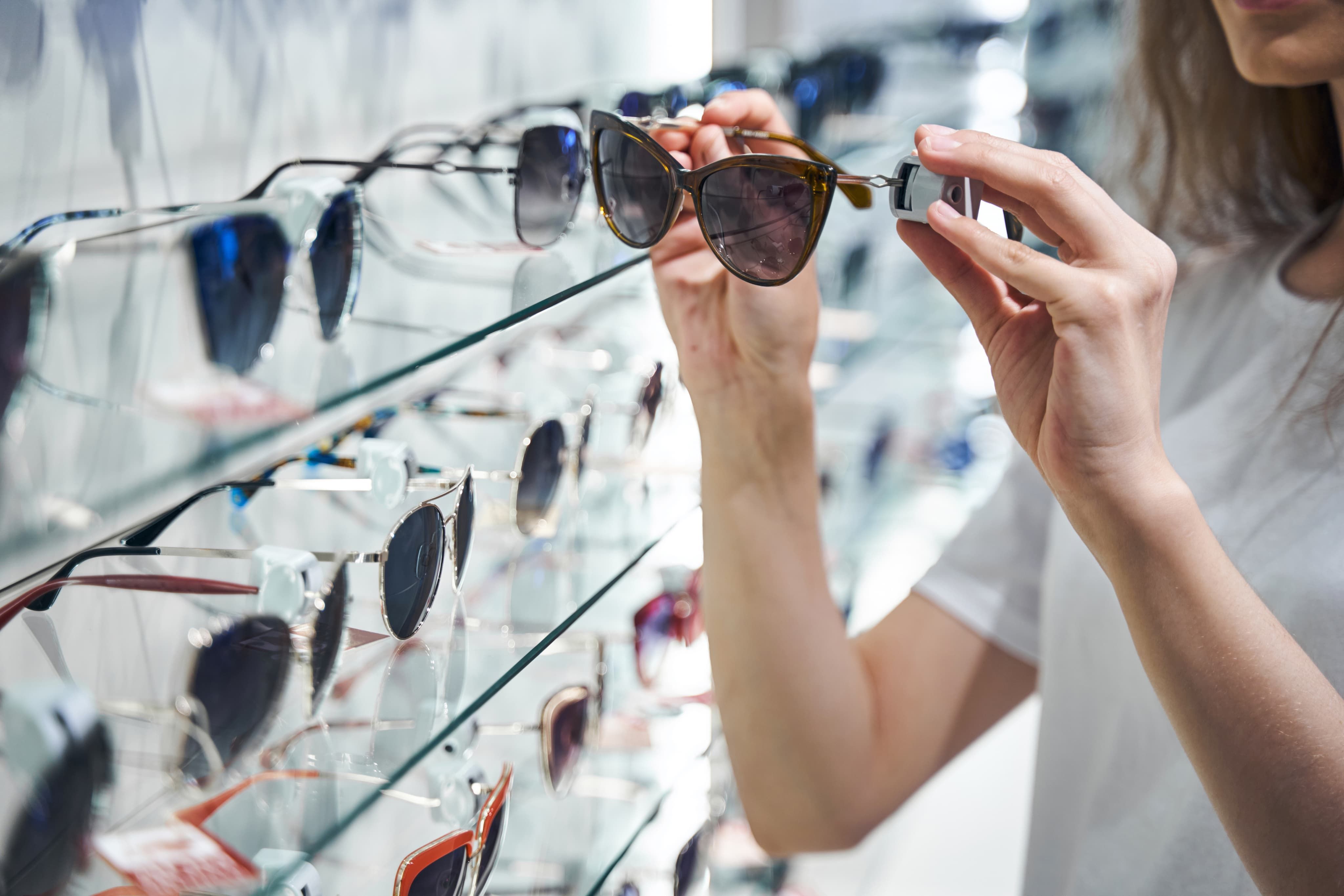 Person picking a cat-eye tortoiseshell pair of sunglasses from a glass display rack in an eyewear store.