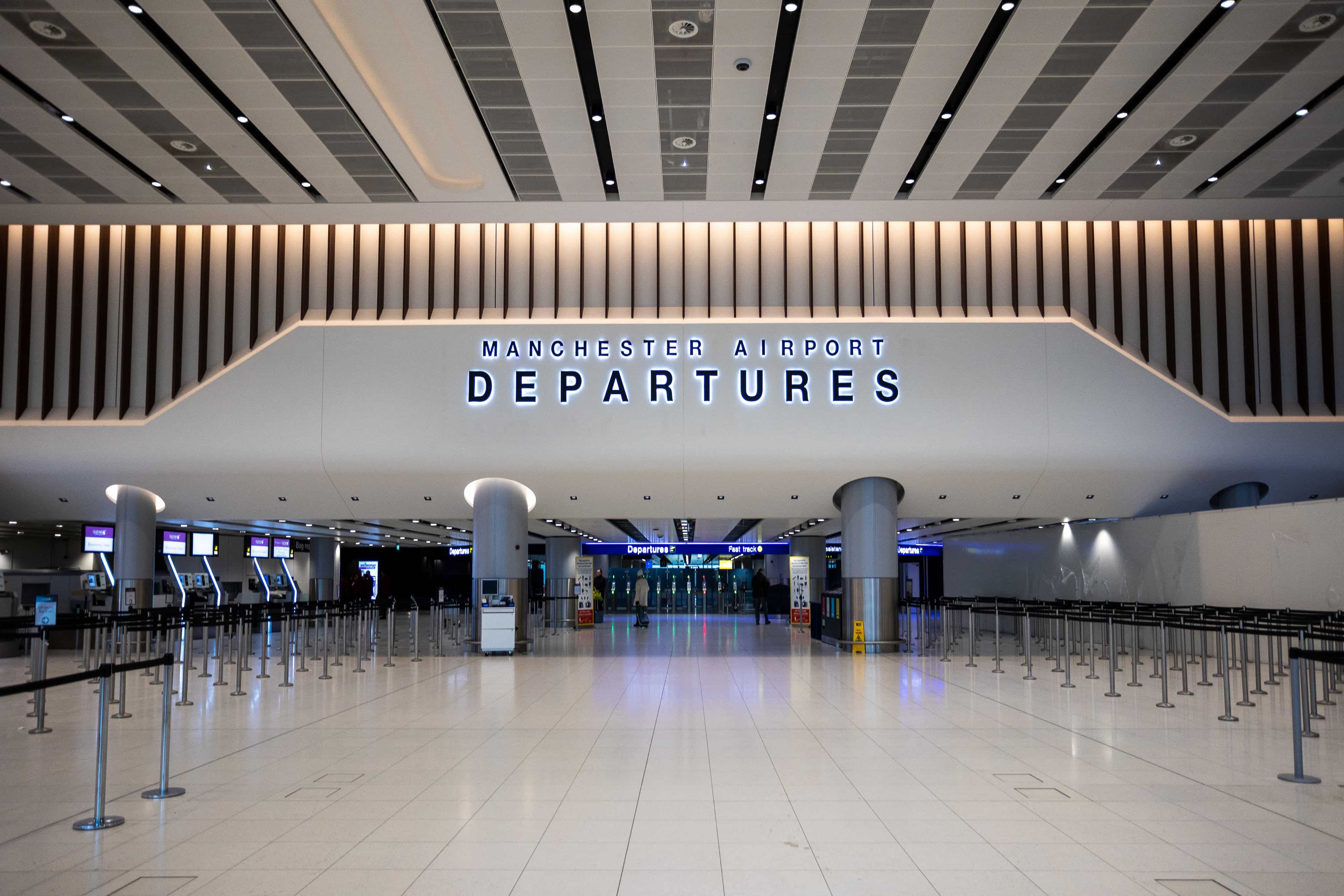 Wide view of Manchester Airport departures hall with modern design, illuminated "Departures" sign, and empty check-in counters.