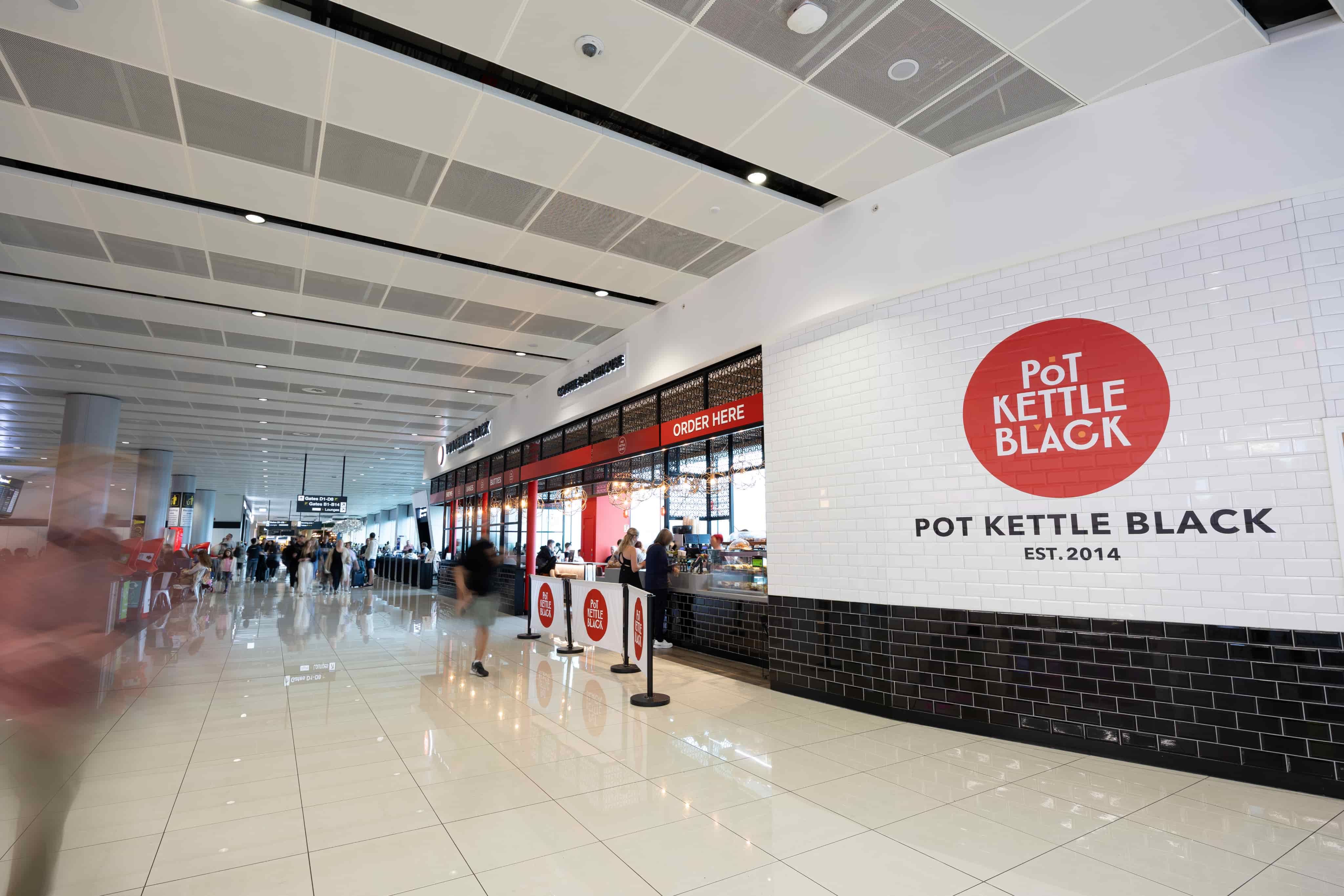 Cafe "Pot Kettle Black" in an airport with a sleek, modern design, featuring a glossy tiled wall and a large red logo.