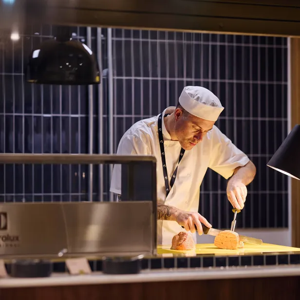 A chef in a white uniform slices meat under warm lighting in a professional kitchen setting.
