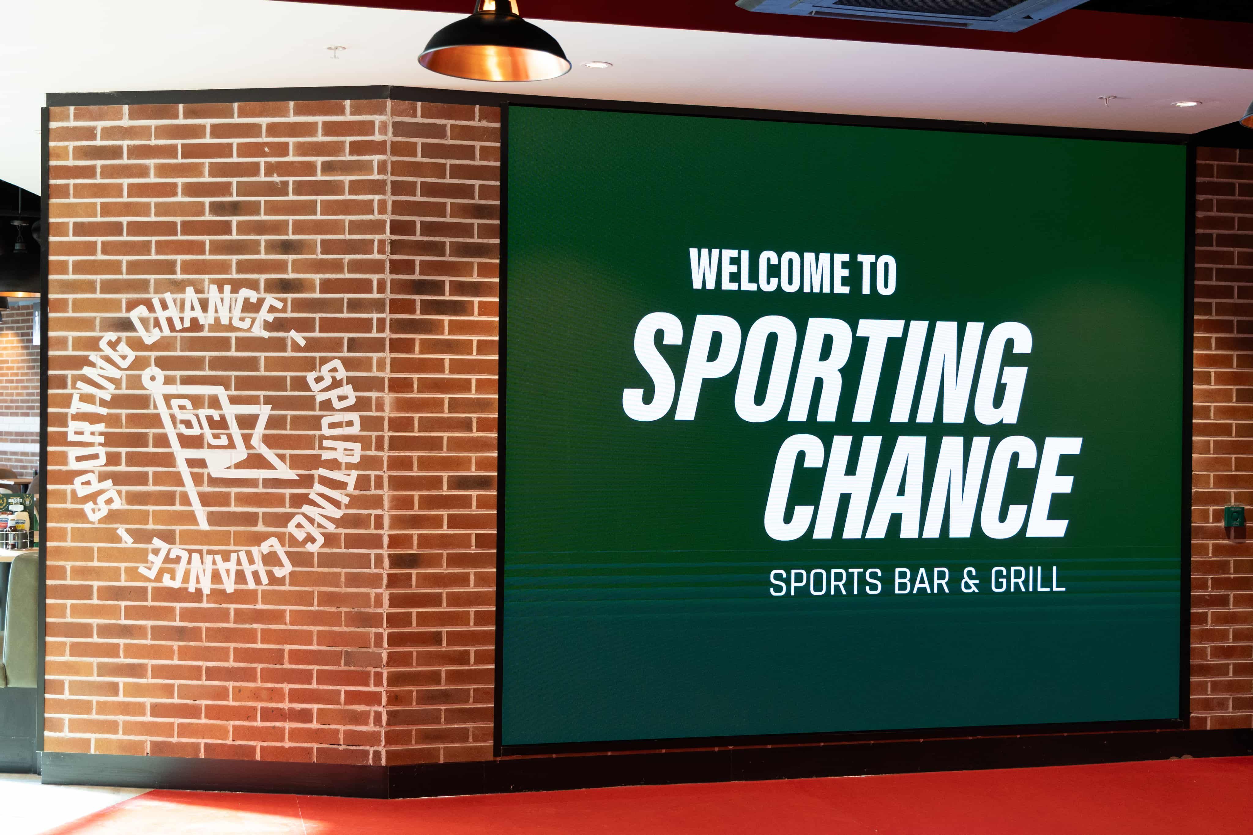 Entrance with a large digital sign reading "Welcome to Sporting Chance Sports Bar & Grill" against a brick wall.