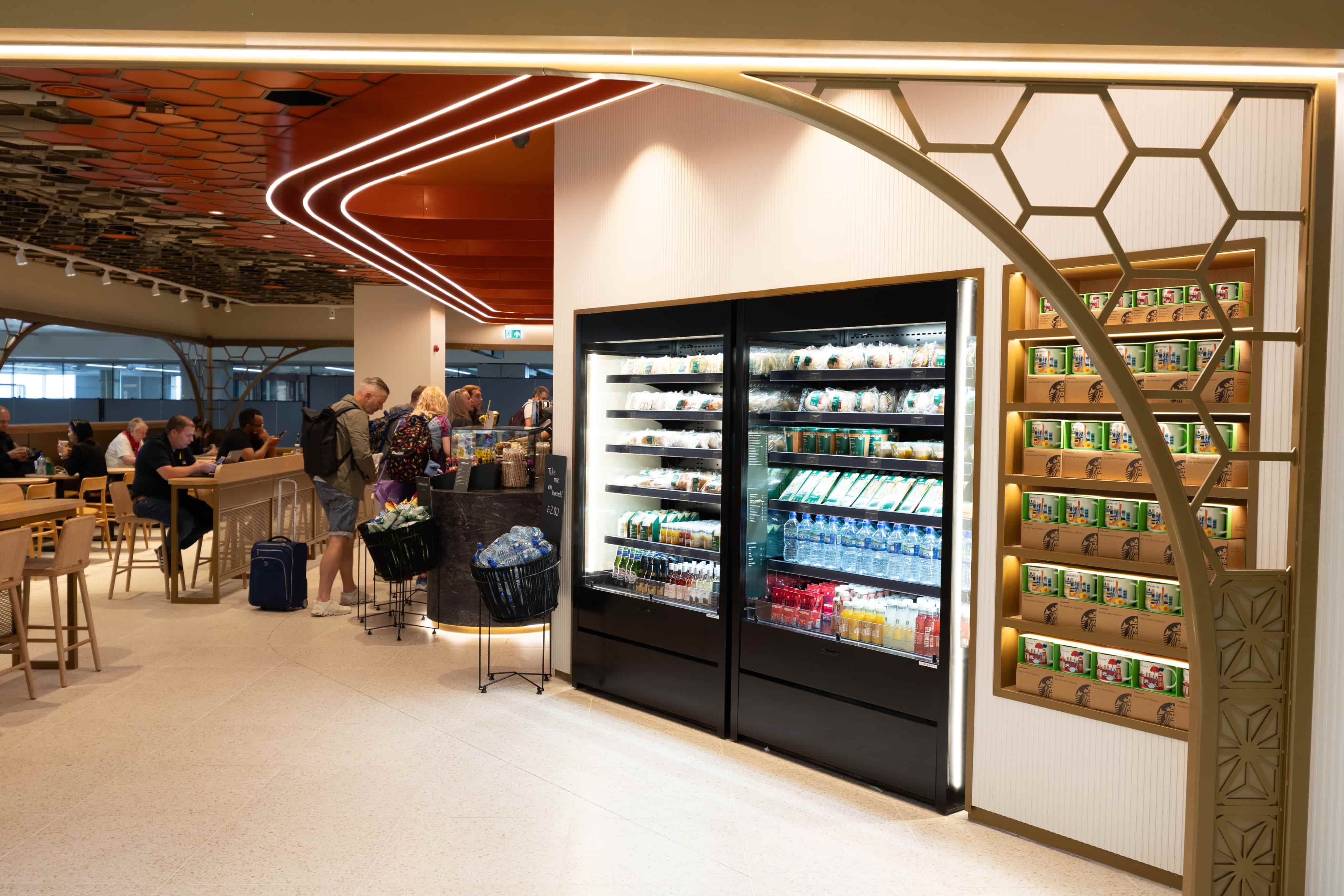 Modern airport café with people ordering at a counter, refrigerated drinks display, snack shelves, and honeycomb ceiling design.