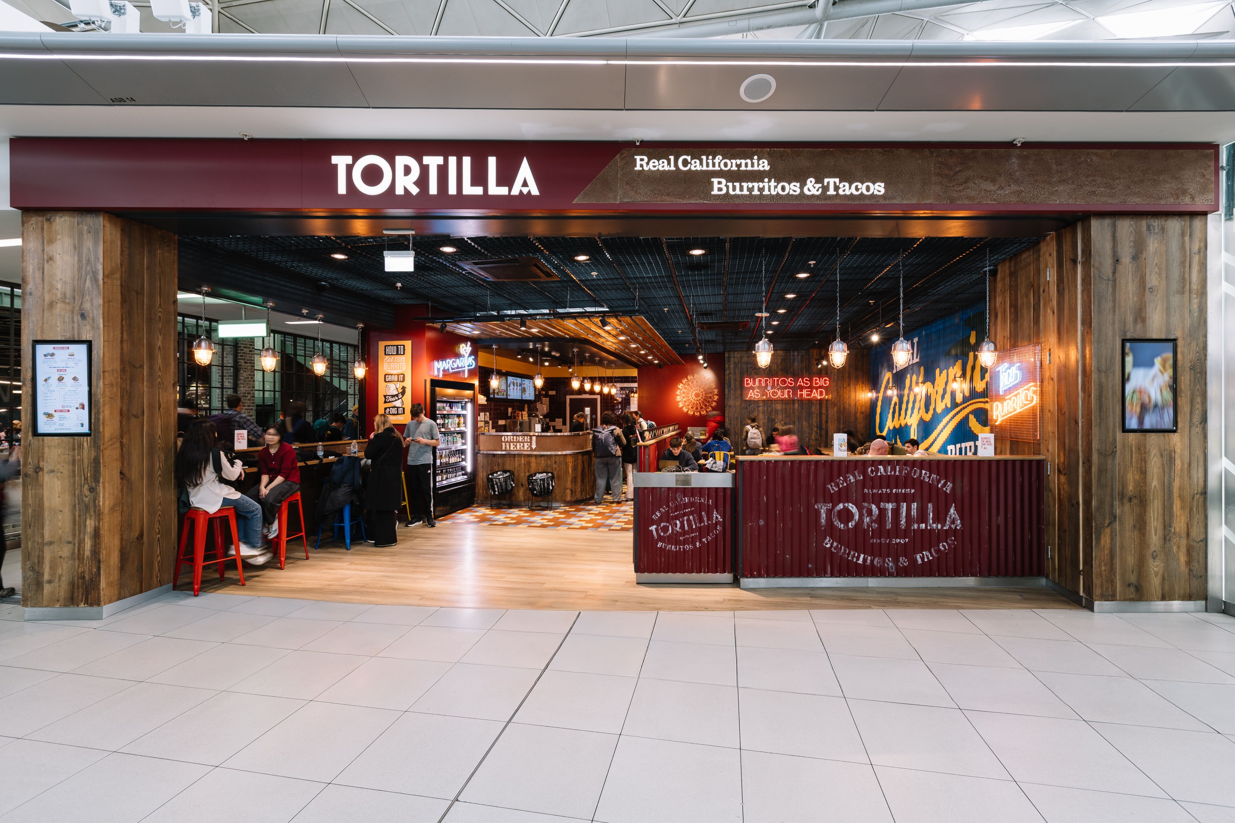 Restaurant entrance labelled "Tortilla" with seating area and counter, offering burritos and tacos under warm hanging lights.