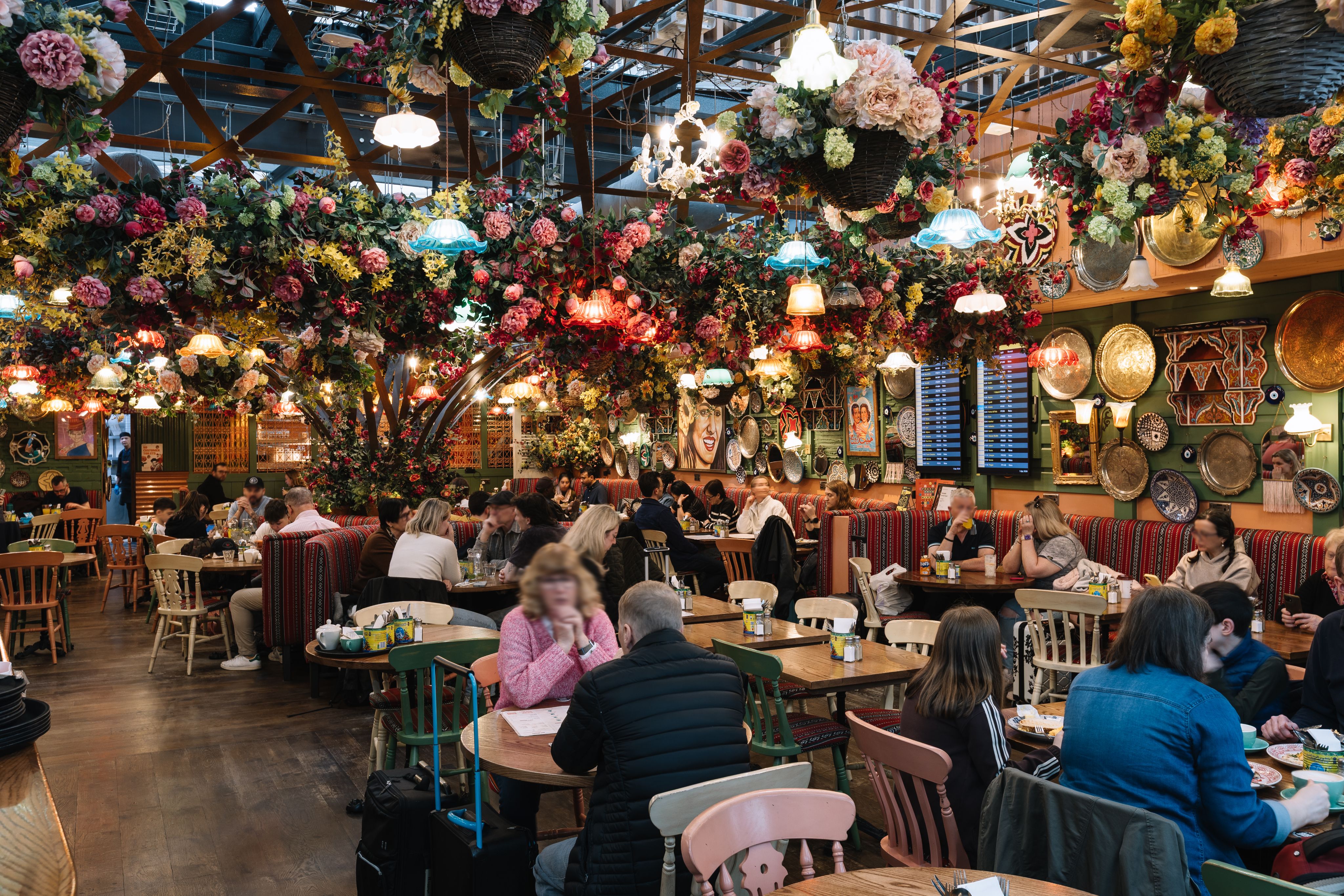 Vibrant restaurant interior with floral décor, colourful hanging lamps, and patrons seated at wooden tables, creating a lively atmosphere.