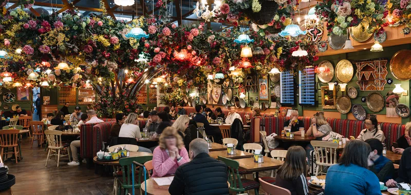 Vibrant restaurant interior with floral décor, colourful hanging lamps, and patrons seated at wooden tables, creating a lively atmosphere.