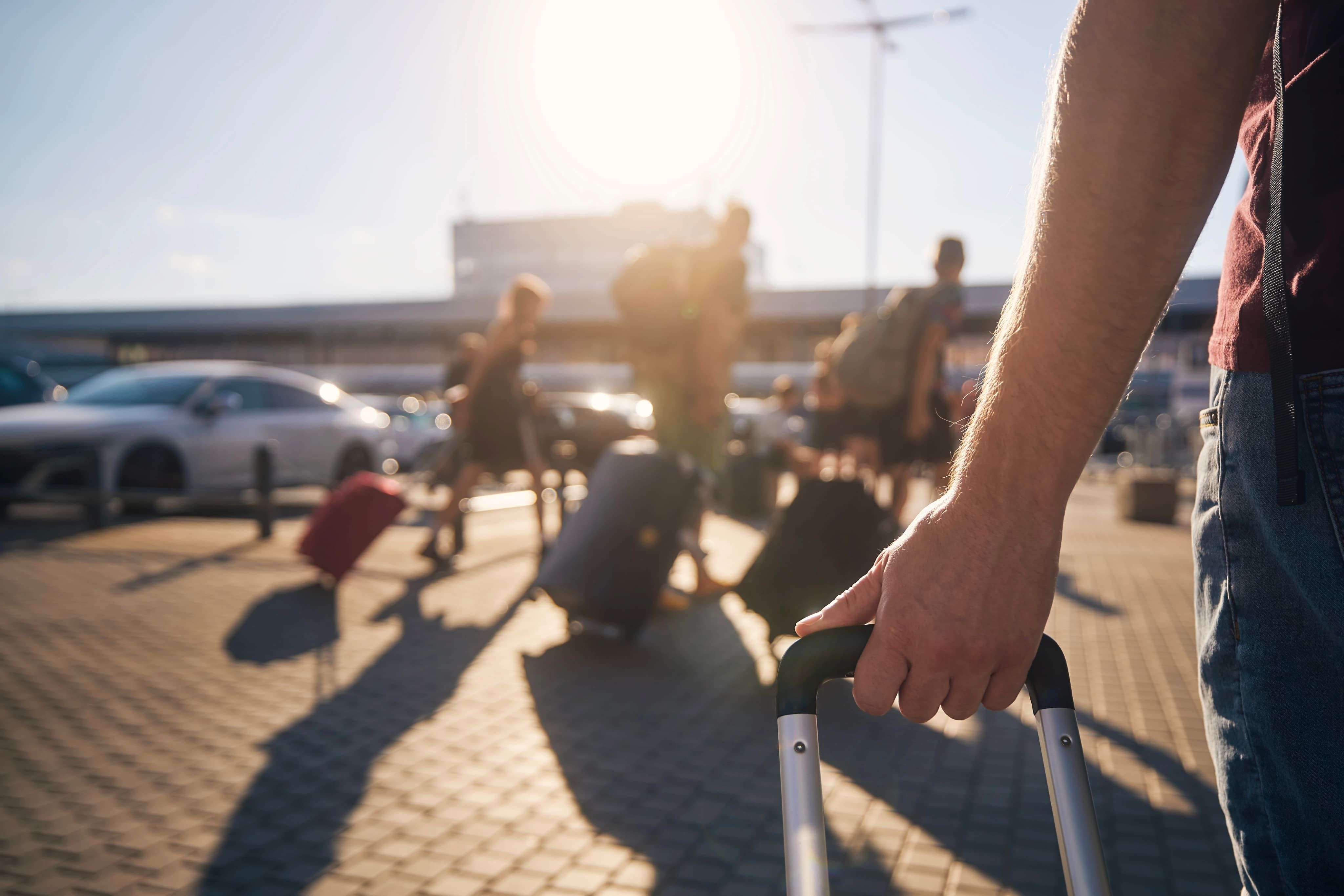 Person pulling a suitcase at a busy outdoor airport area, with people and luggage silhouetted against a bright, sunny sky.