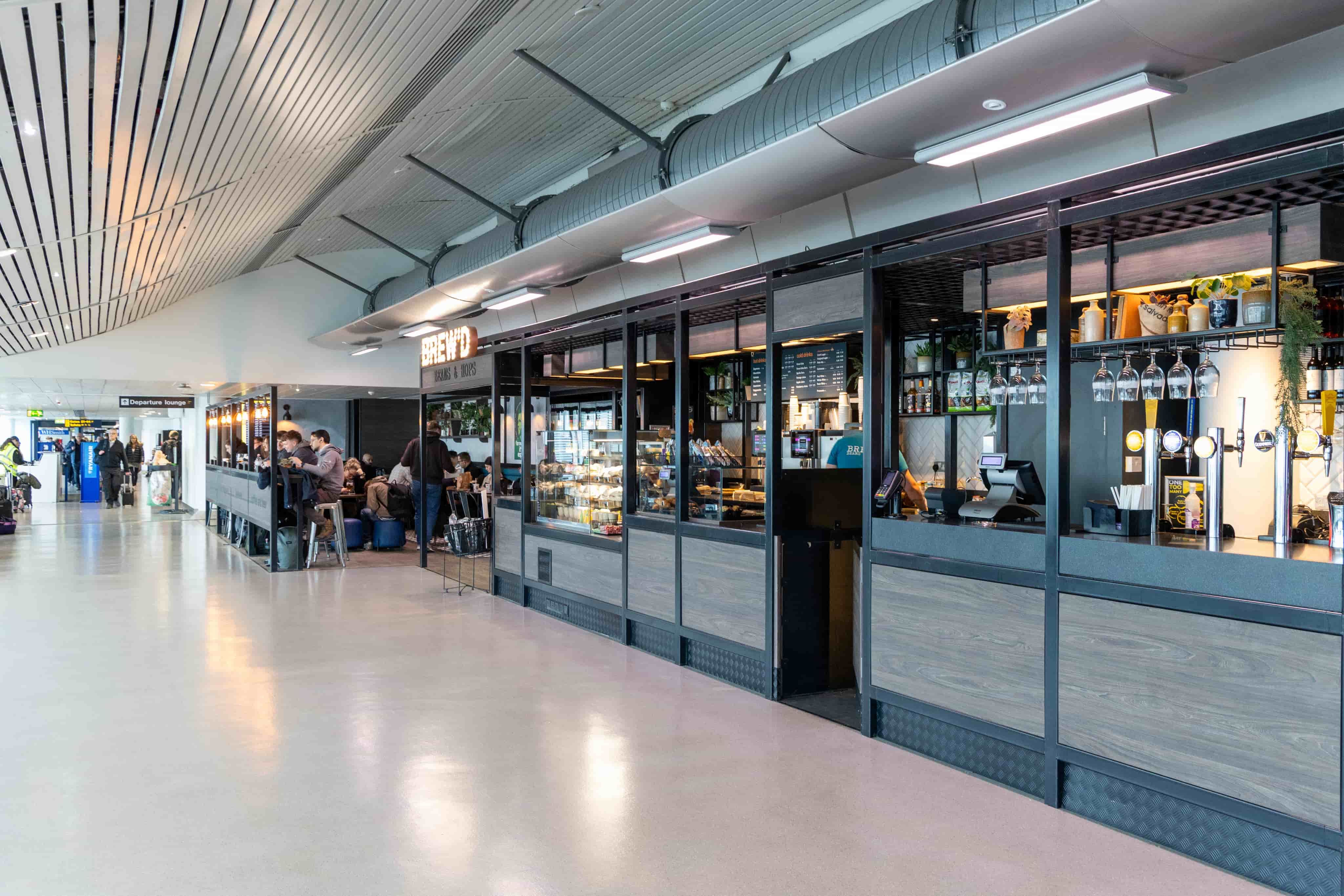 Modern airport café with glass display case, people sitting inside. Overhead lights and sleek design elements complete the contemporary look.