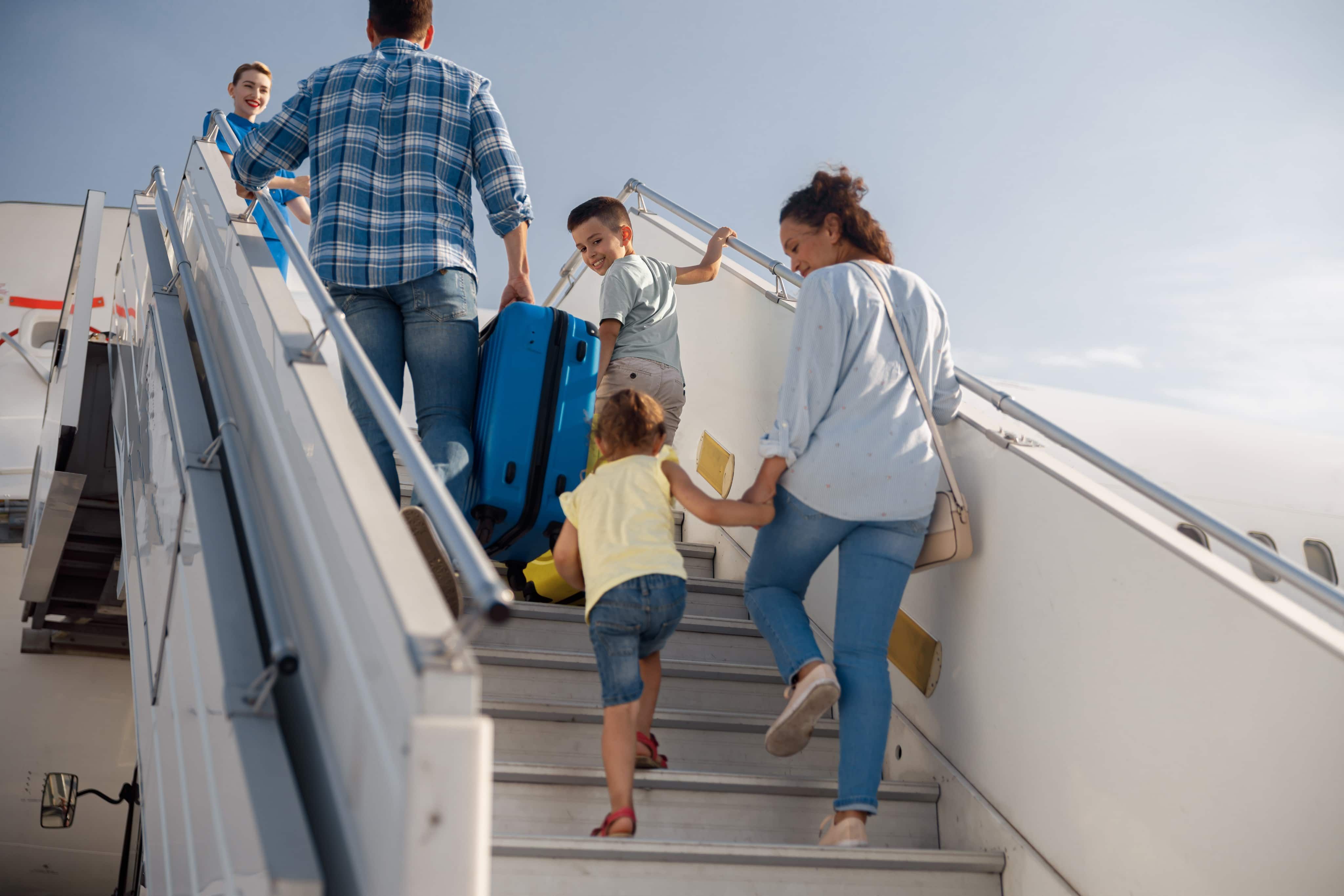 A family, including two children, climbs airplane stairs, carrying luggage and boarding passes, under a clear blue sky.