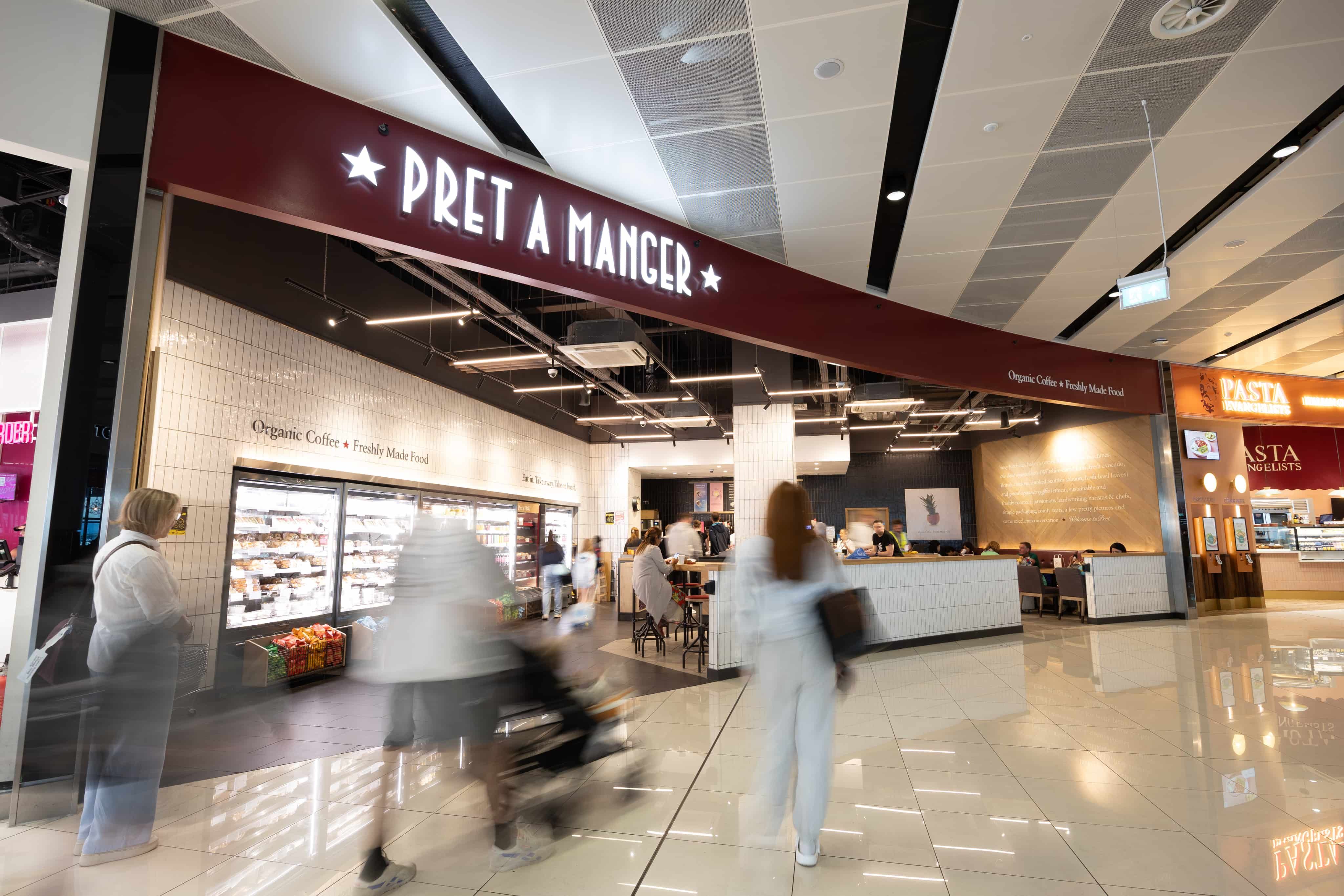 People walking in a spacious shopping area with a Pret a Manger cafe. Some patrons sit at tables; others browse shelves under bright lighting.
