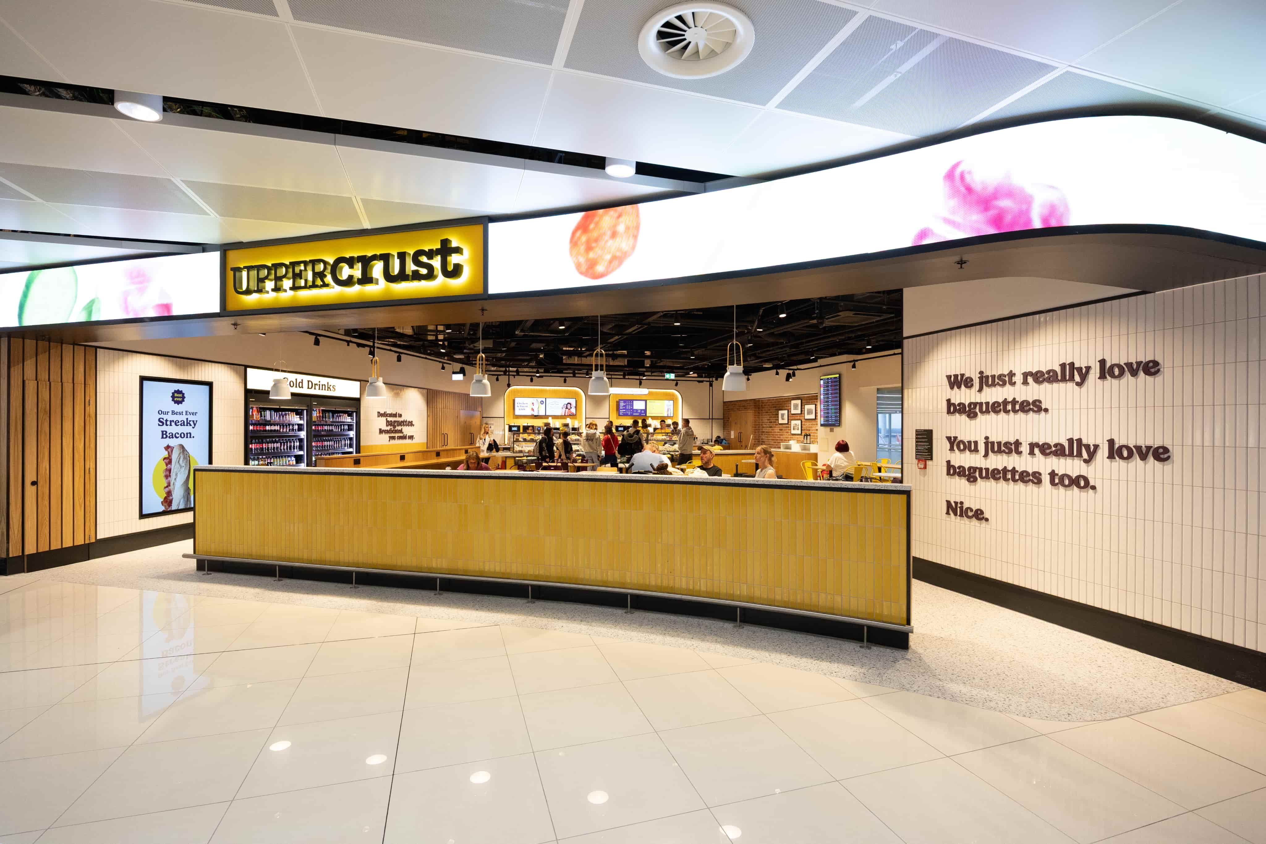 A modern Upper Crust café with a yellow and white theme, diners inside, and wall text about loving baguettes.
