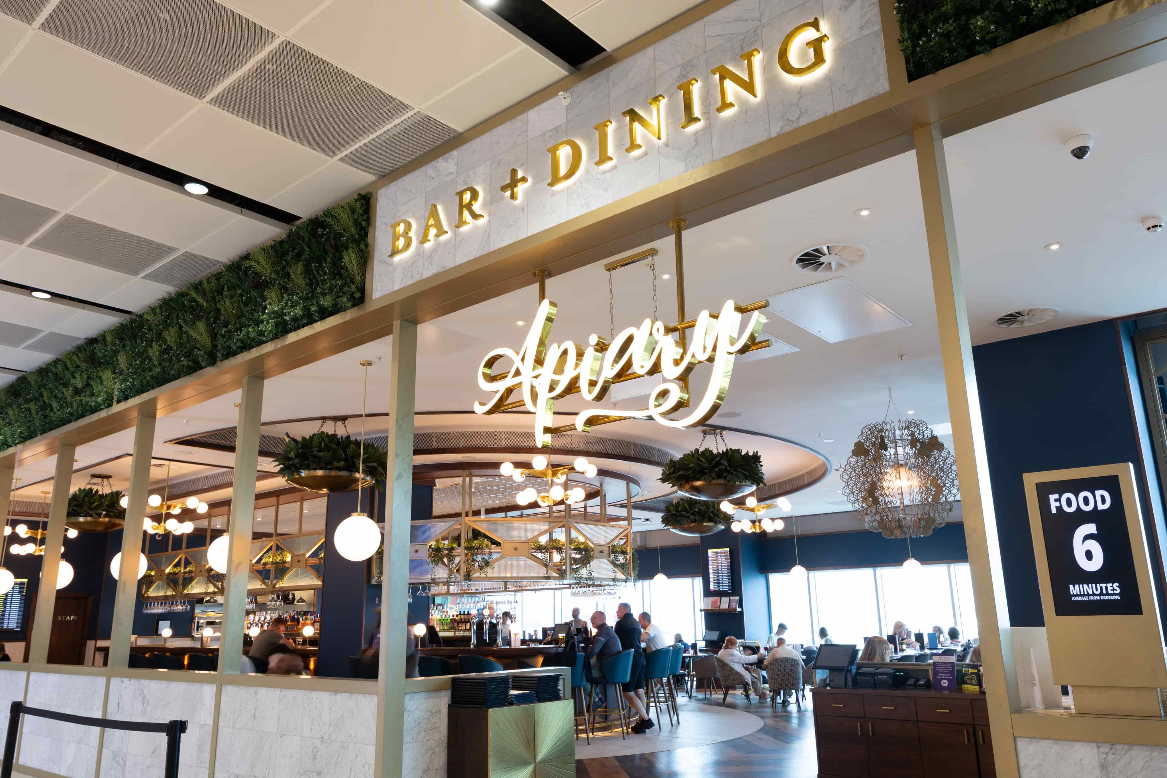 Modern restaurant interior with a sign reading "Bar + Dining" and "Apron." Patrons seated inside, surrounded by elegant lighting and decor.
